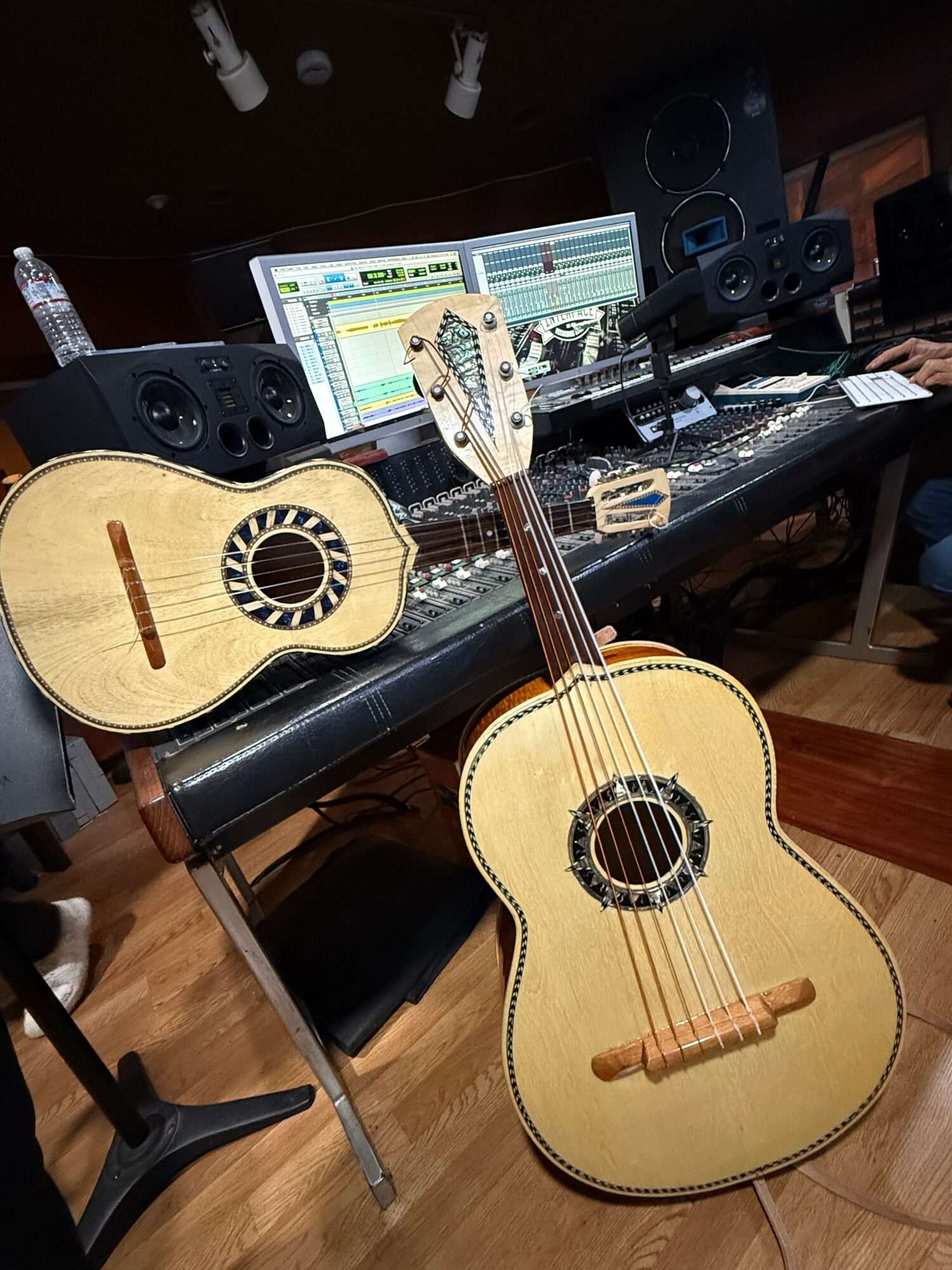 Two acoustic guitars on a desk with a computer monitor, keyboard, and audio equipment in the background.