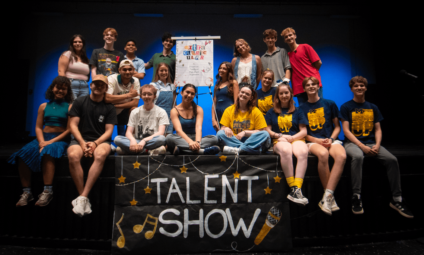 Group of children and teenagers on stage behind a talent show banner, some sitting and some standing, with a colorful poster behind them.