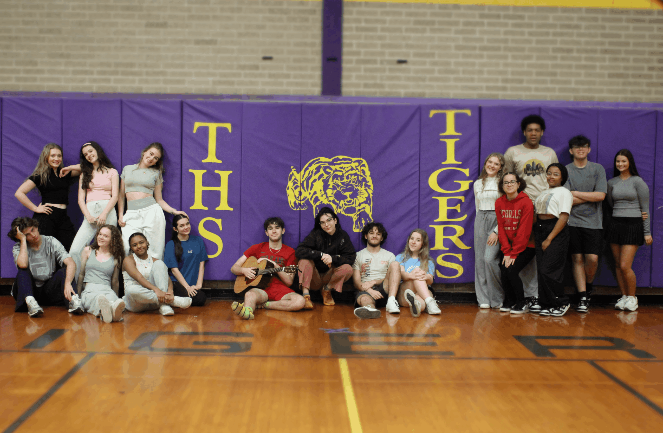 Group of children and teenagers in a gymnasium with purple and yellow tiger-themed backdrop, some sitting and some standing.