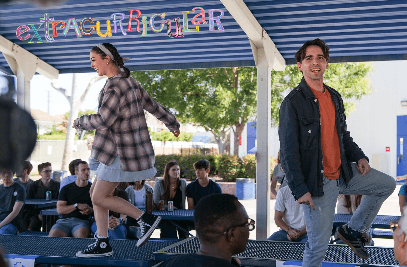 Two students on a stage, one jumping and the other smiling, with a group of students seated behind them outdoors.