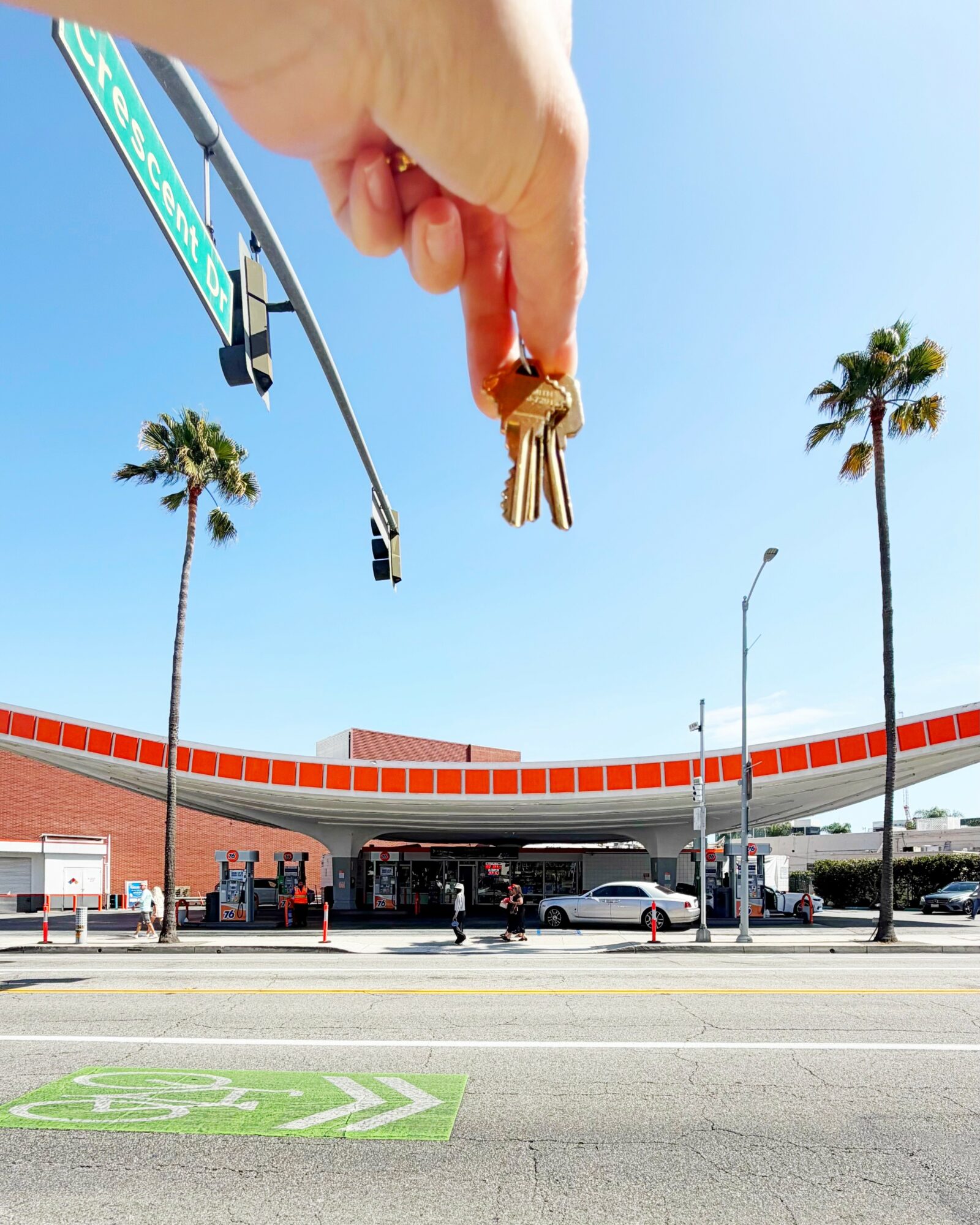 Hand holding keys above a street with palm trees and a building, sky is clear and blue.