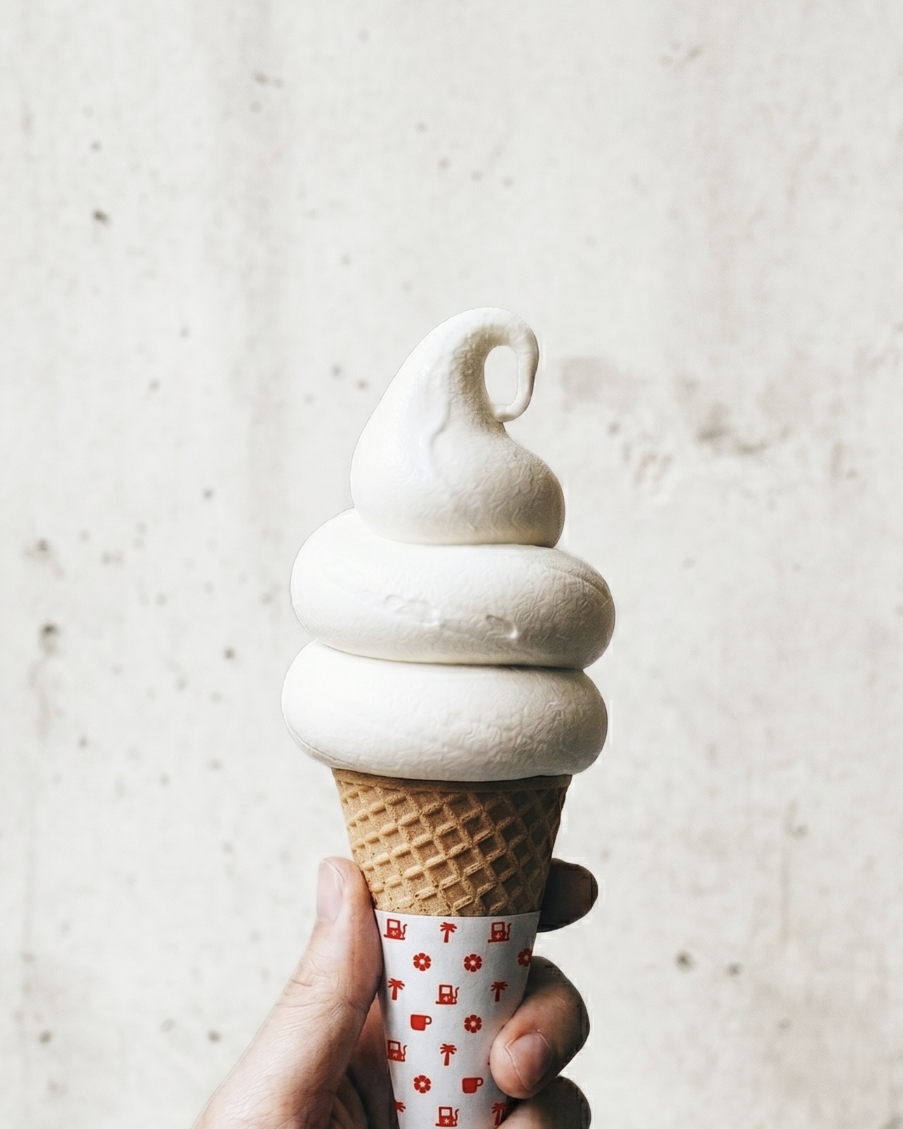 Hand holding an ice cream cone with three scoops of white ice cream against a plain background.