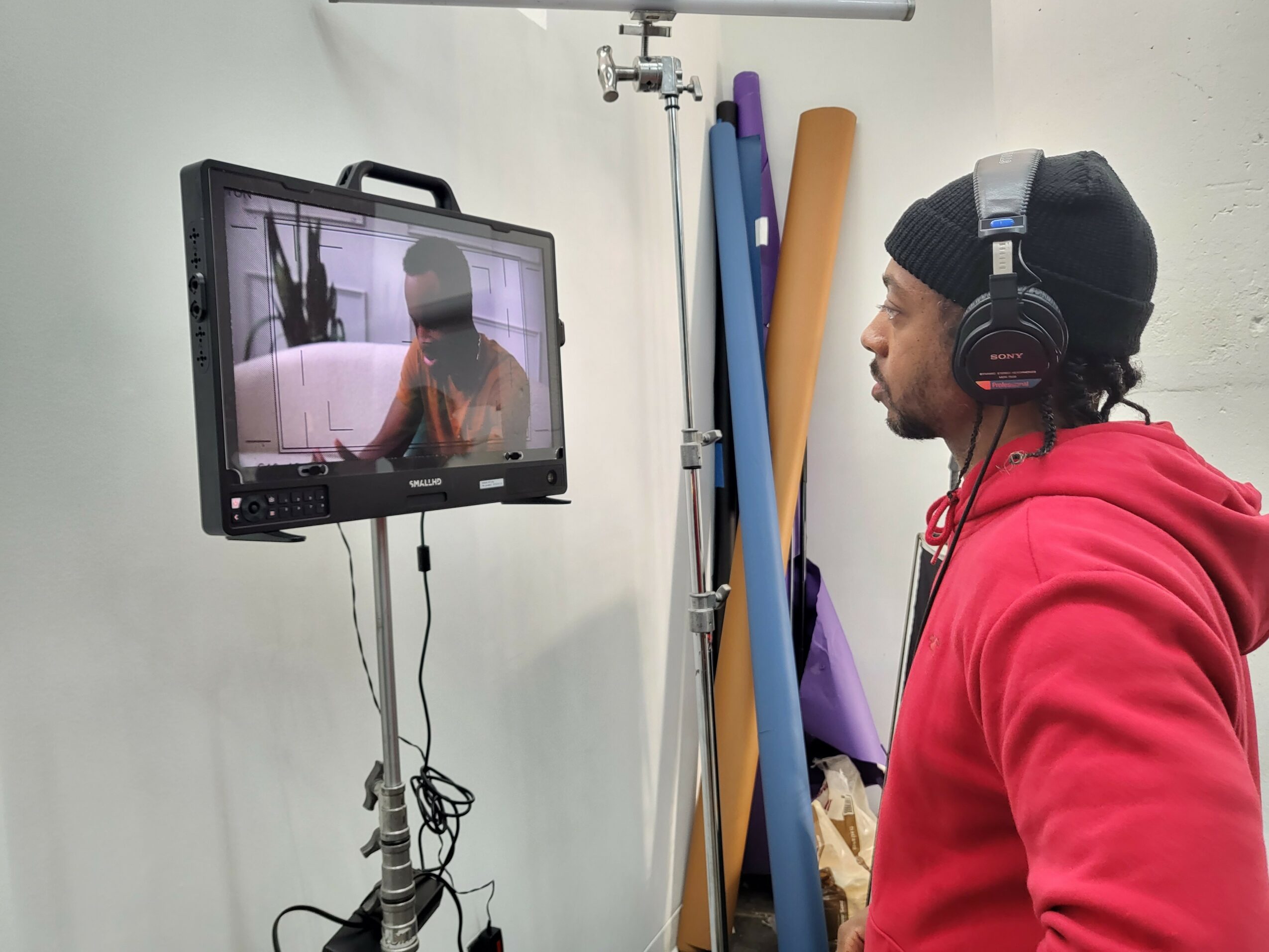 Man wearing headphones and a black beanie watches a monitor showing a person in a studio setting.