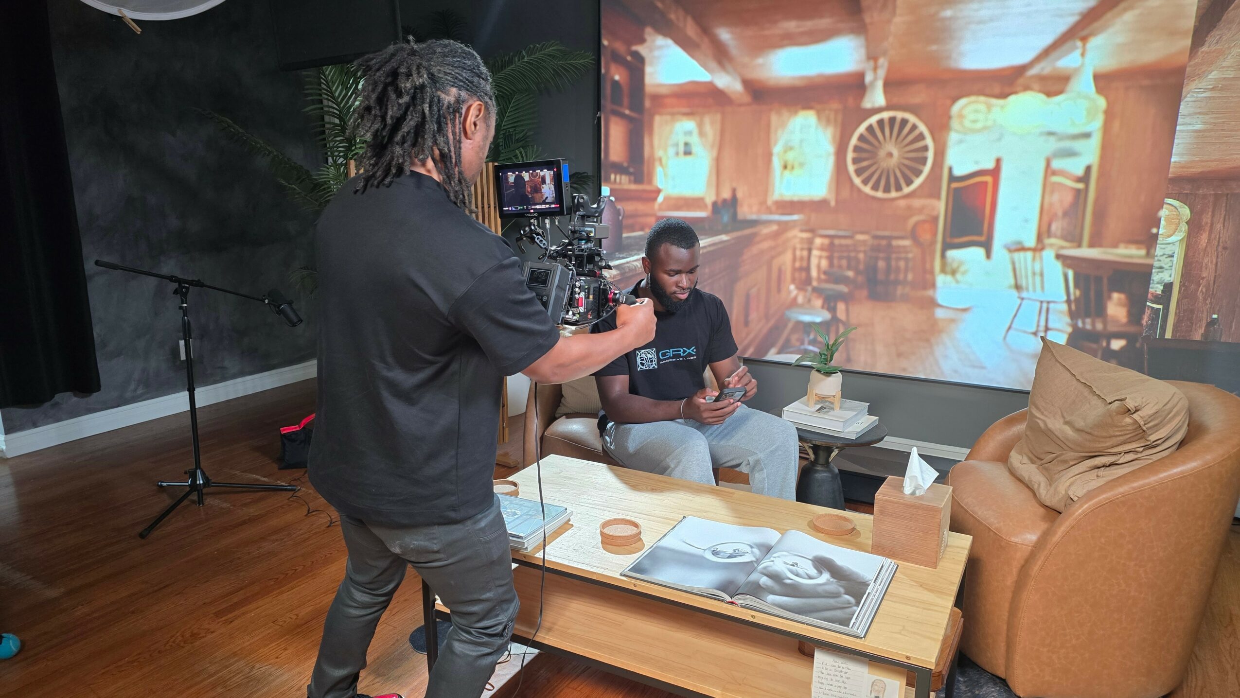 Person filming a child sitting on a couch in a room with a large screen showing a library scene, wooden floor, and furniture.
