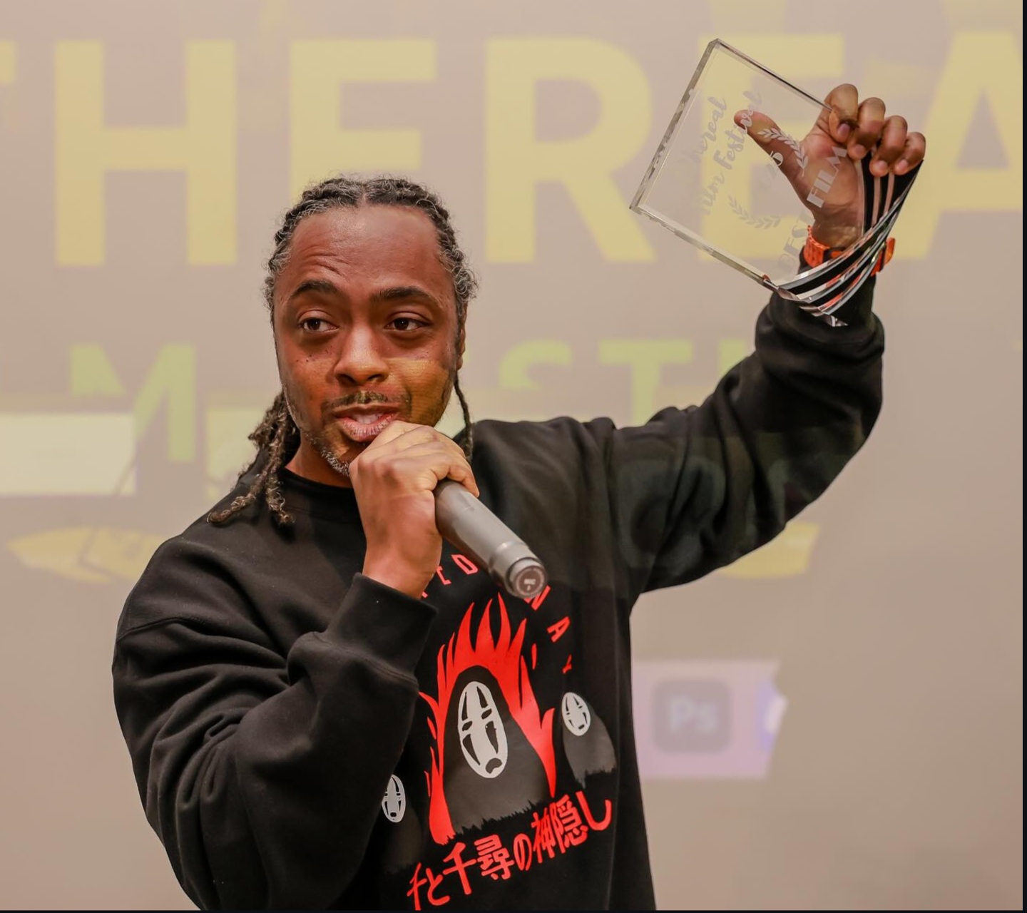 Man with dreadlocks holding a microphone and a transparent award, speaking or presenting, background with large yellow text.