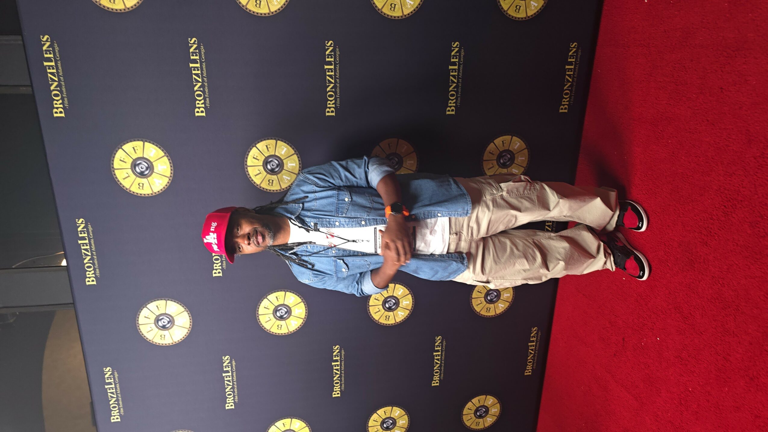 Person standing on red carpet in front of a black backdrop with gold logos, wearing a red cap, denim jacket, and beige pants.