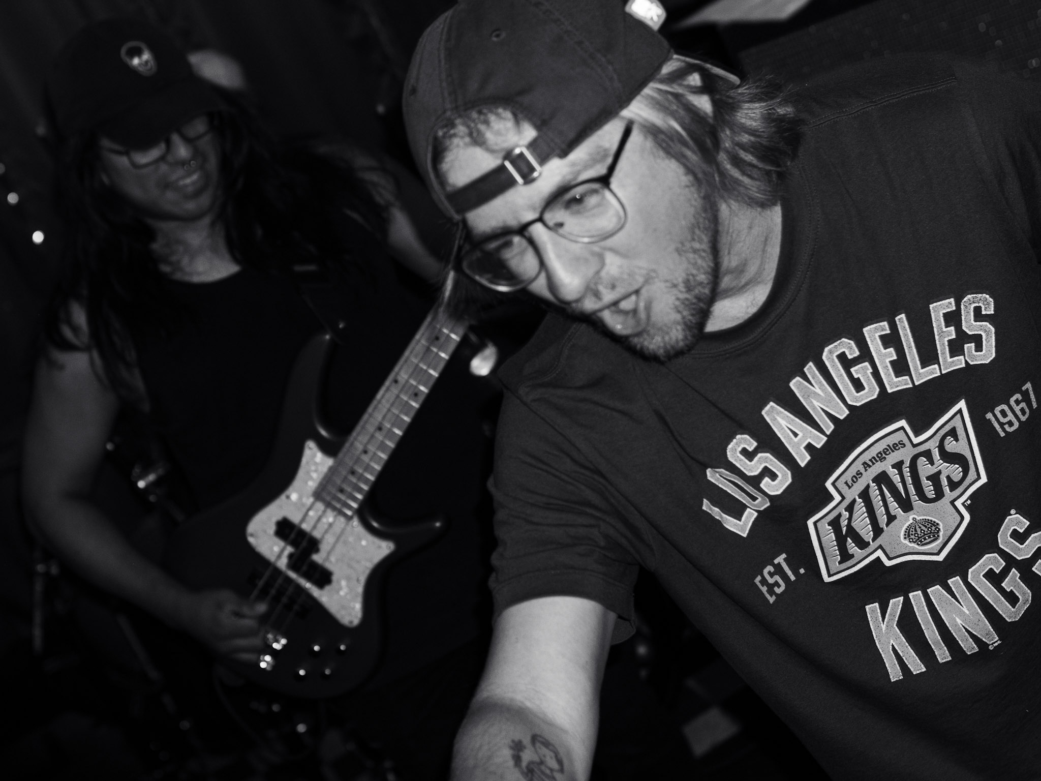 Two people, one playing guitar, in a dark setting, with one wearing glasses and a cap, and the other with long hair.