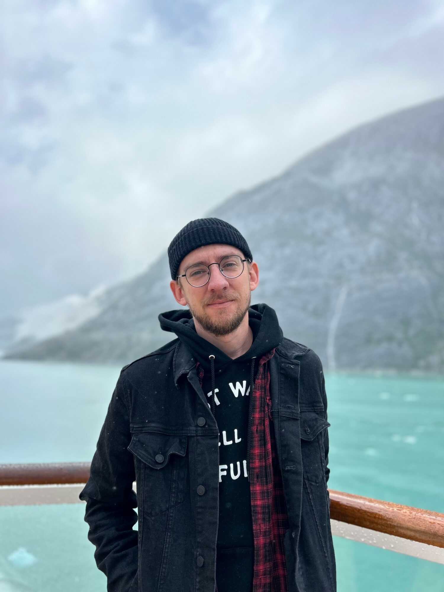 Young man with glasses and a black beanie stands outdoors near water with mountains in the background.