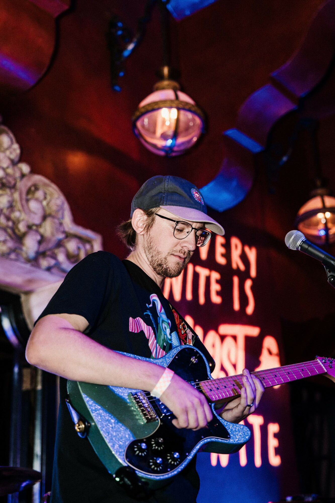 A man with glasses and a cap plays an electric guitar on stage with a red background and decorative lights.