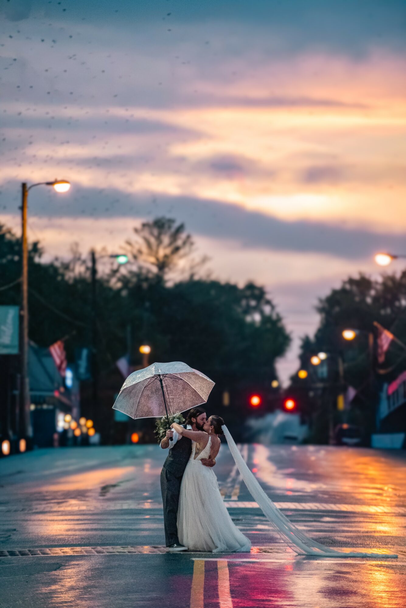 Couple with umbrella standing in the rain on a street at sunset, with streetlights and trees in the background.