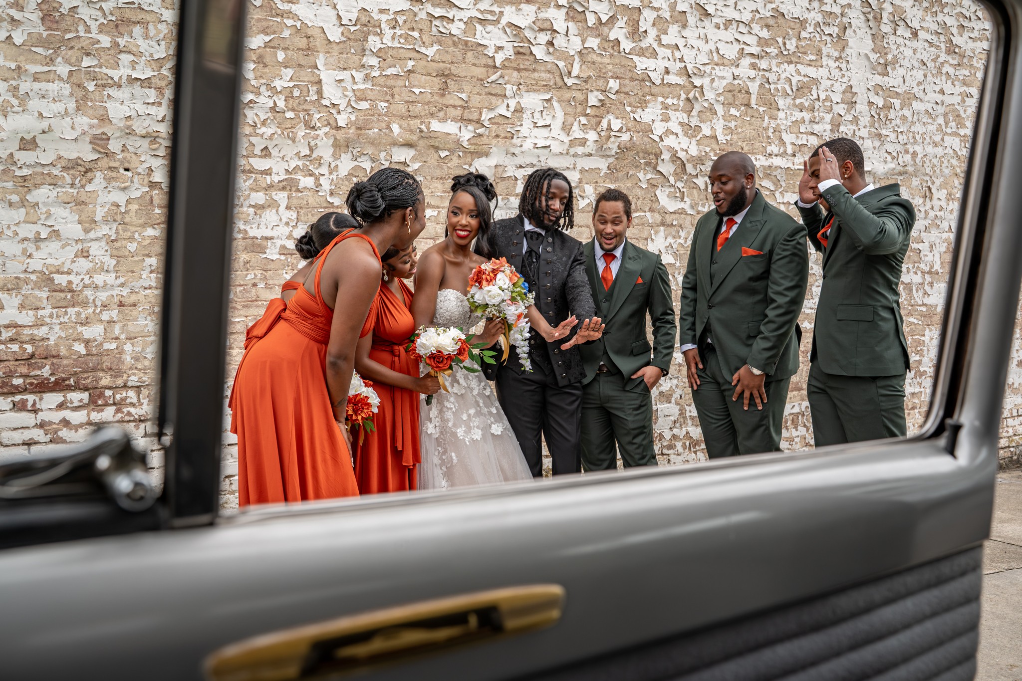 Group of people dressed in formal attire, standing outdoors against a brick wall, with a bride holding a bouquet, smiling and talking.