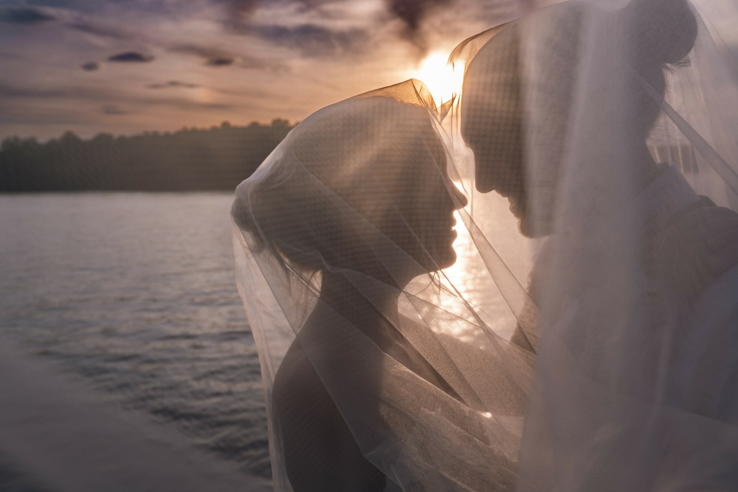 Silhouette of a couple close together with a sunset over water in the background, their faces near each other, partially obscured by a veil.