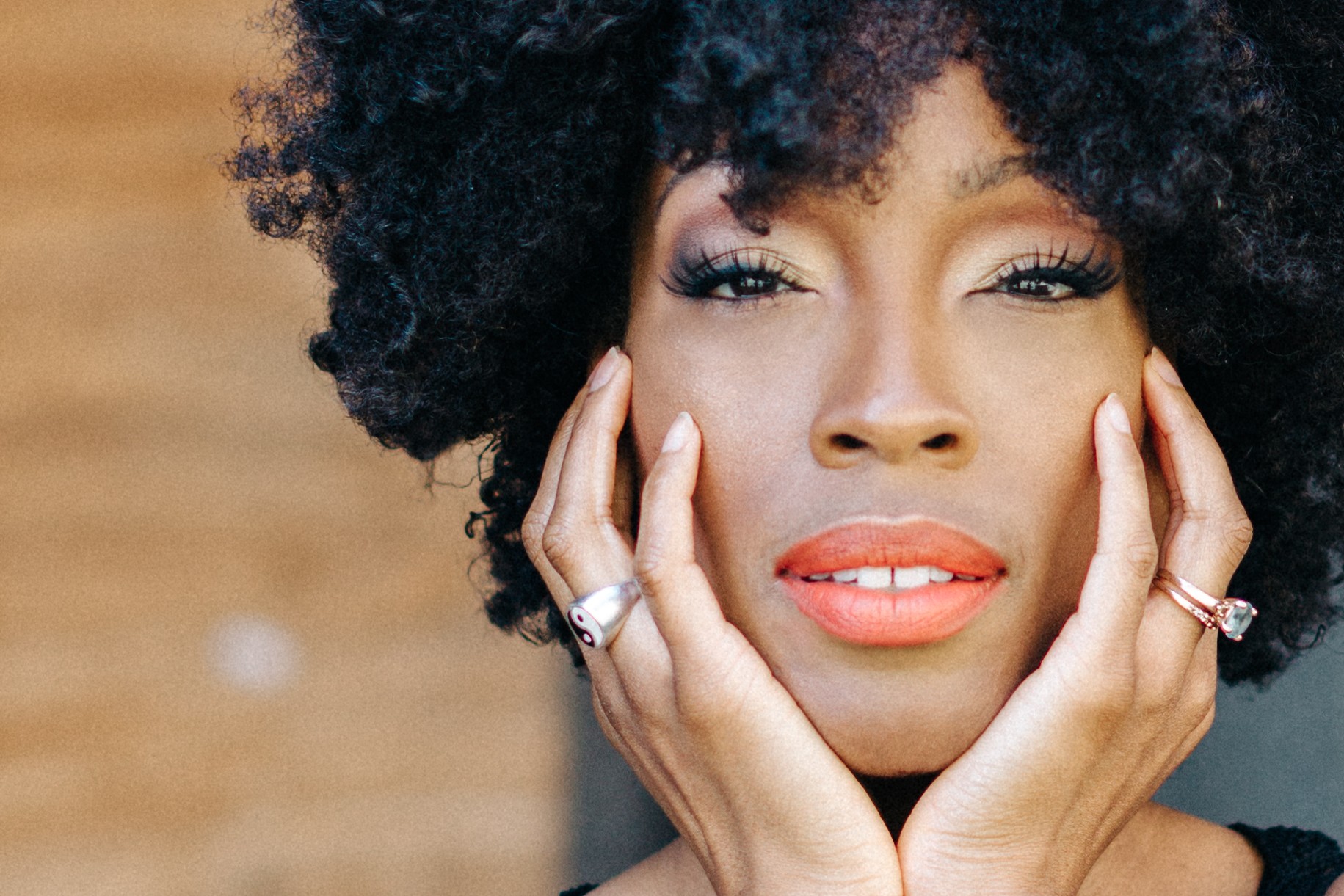 Close-up of a woman with curly hair, touching her face with both hands, wearing rings, and looking at the camera.
