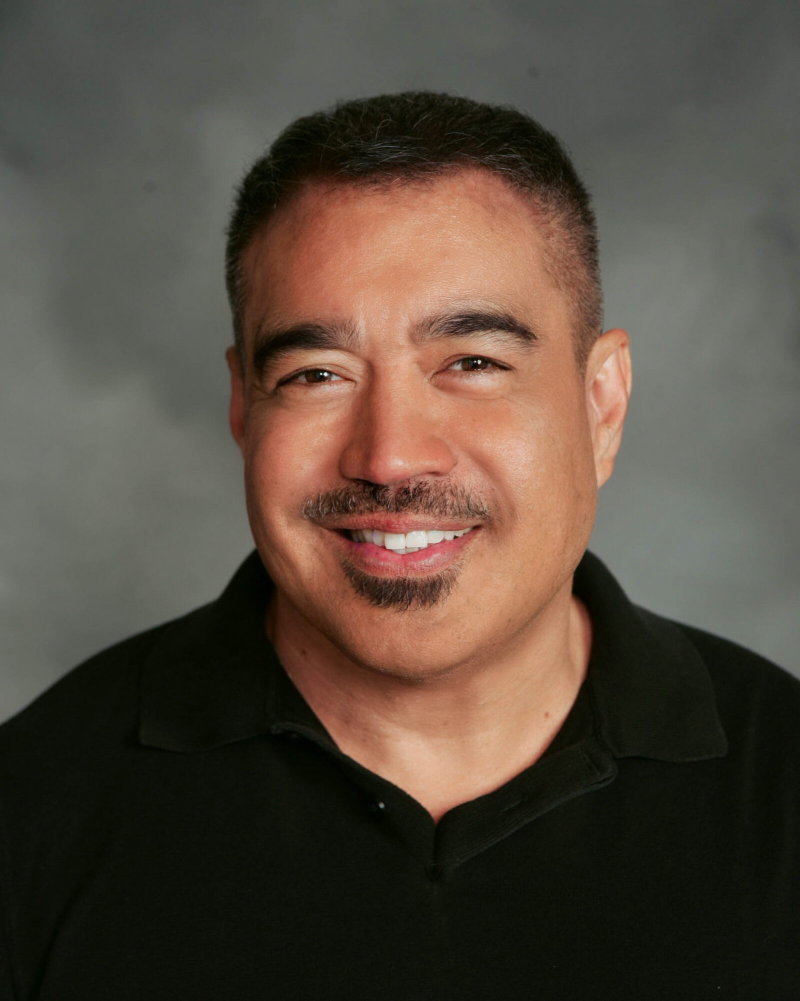 A smiling man with short dark hair and a goatee, wearing a black collared shirt, against a gray background.
