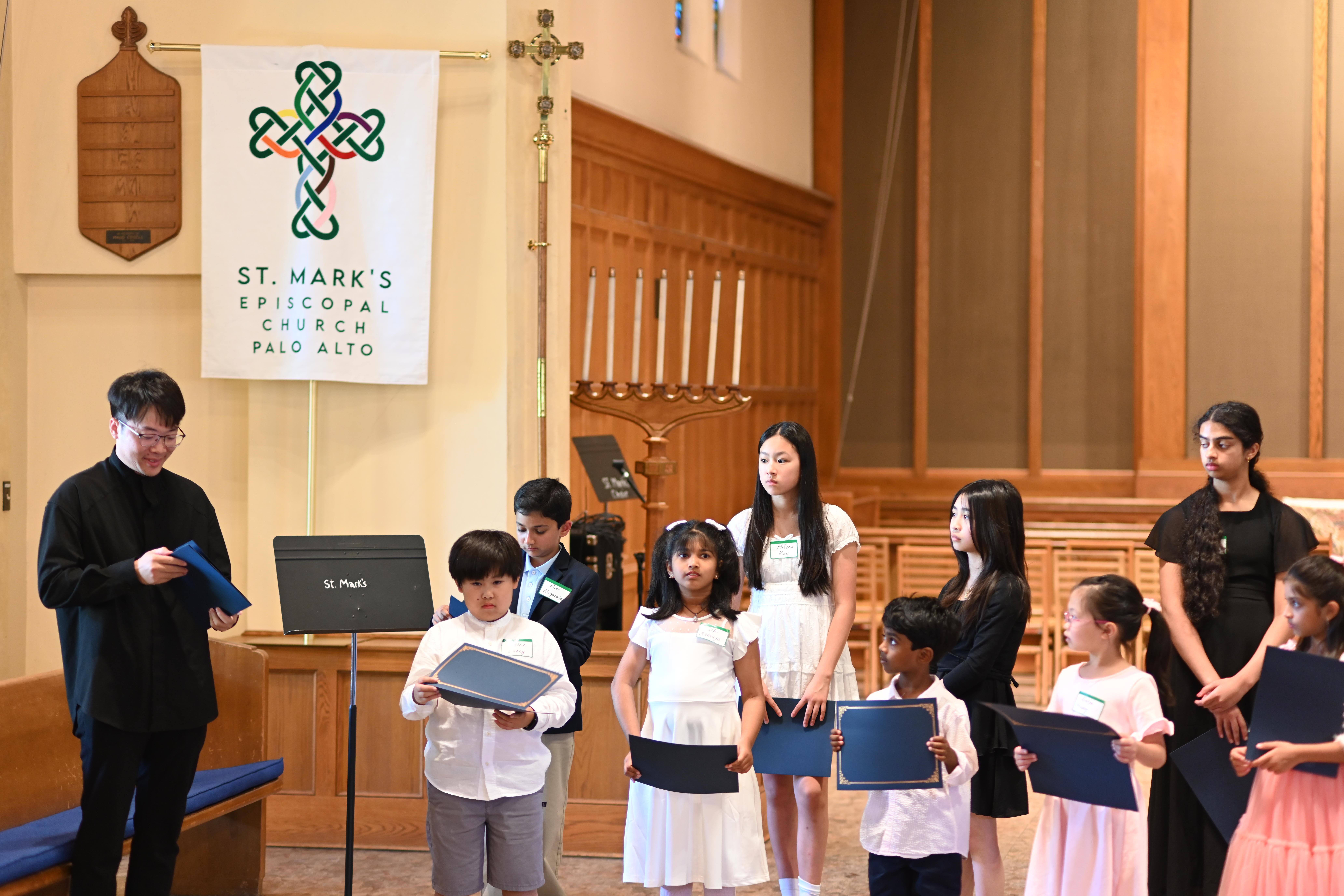 Group of children and adults in a church, some holding books, with a priest and a banner in the background.