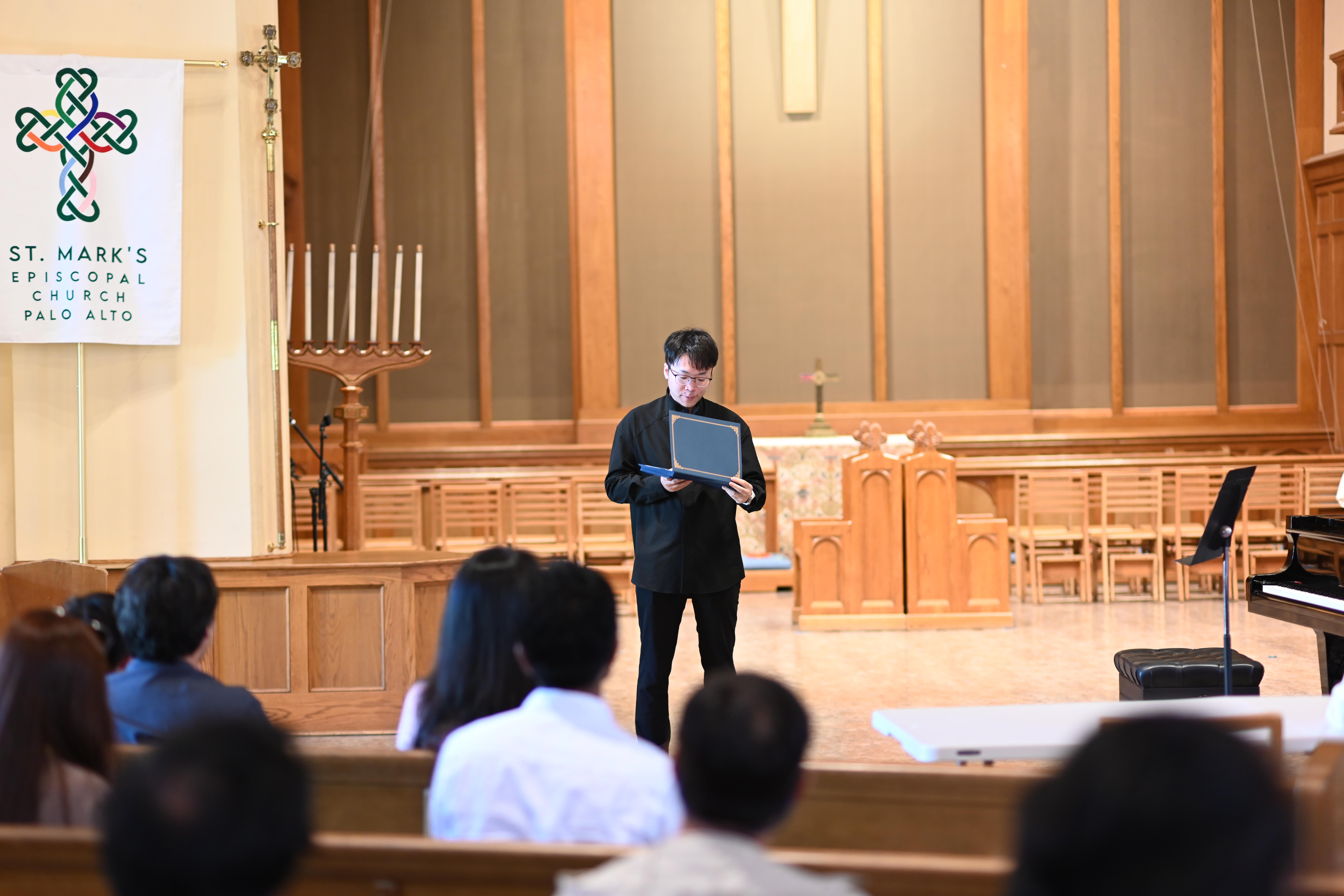 Person standing on stage holding a book or folder, addressing an audience in a church or auditorium, with wooden paneling and a cross in the background.