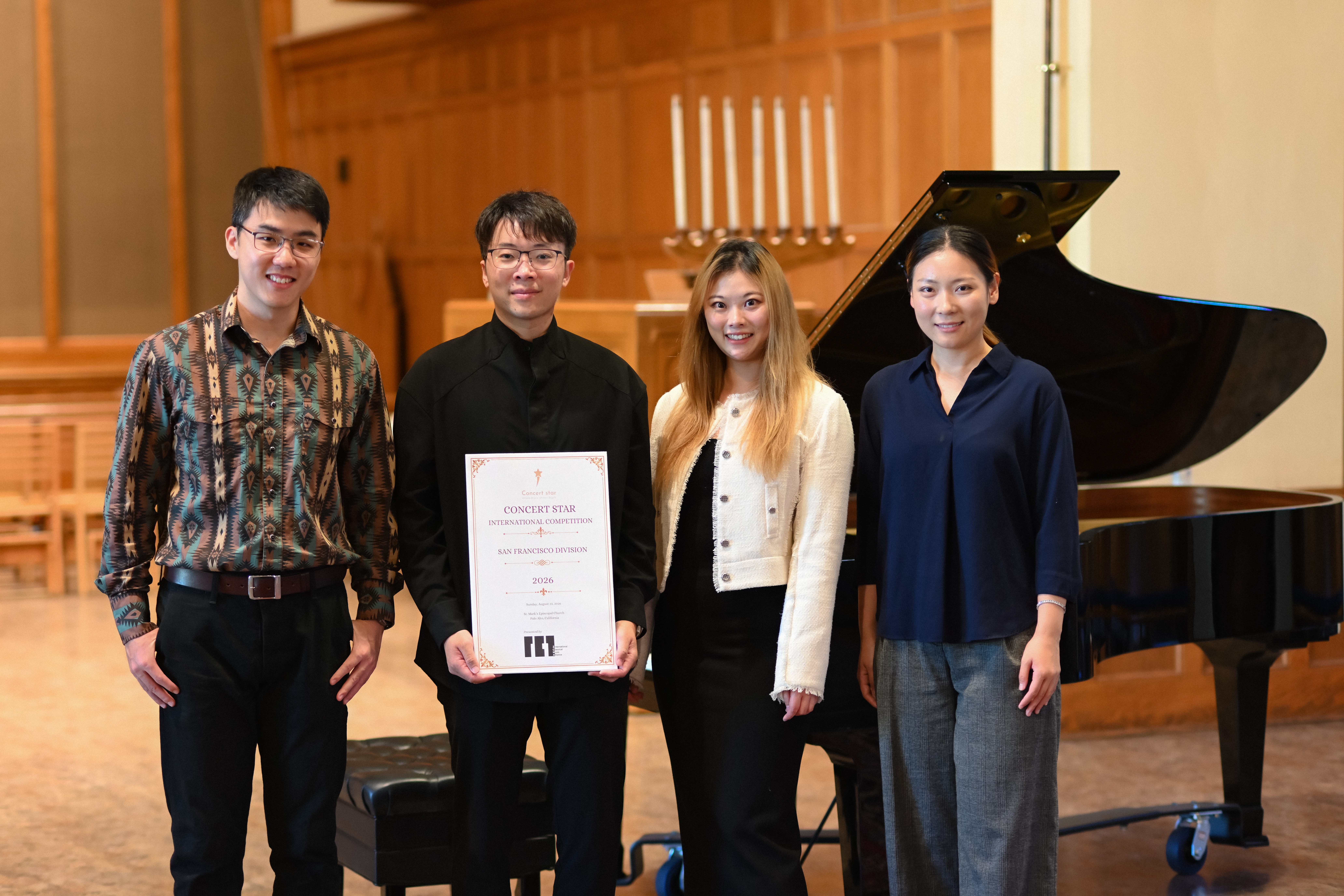 Four people standing on stage in front of a grand piano, smiling, with wooden paneling in the background.