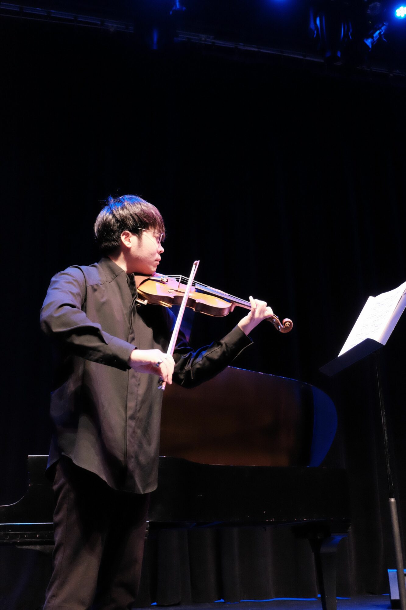 Person in formal attire holding a violin and bow, standing on a stage with dark background.