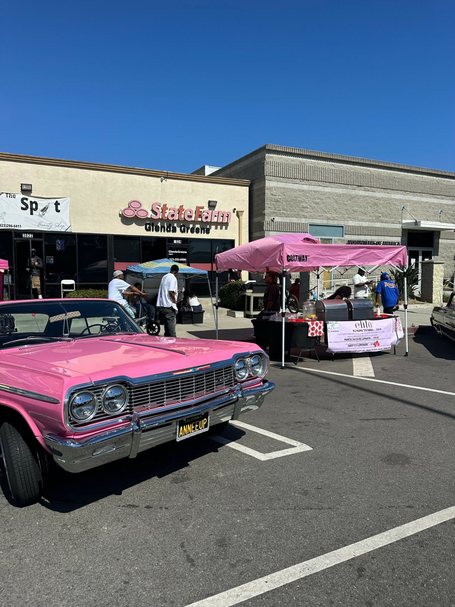 Pink vintage convertible car parked in a lot with a pink tent and people nearby, storefront in background.