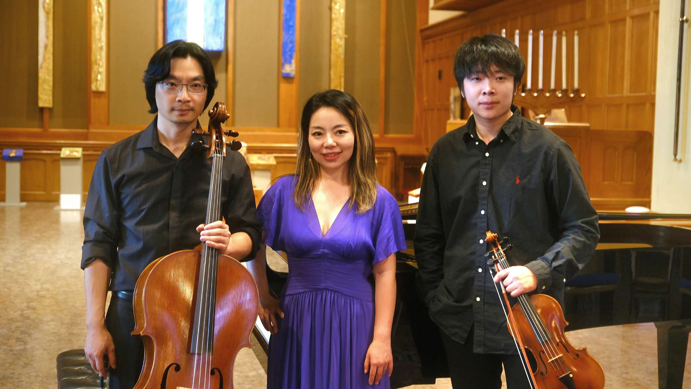 Three people standing in a music hall, two holding violins, one in the middle wearing a purple dress.