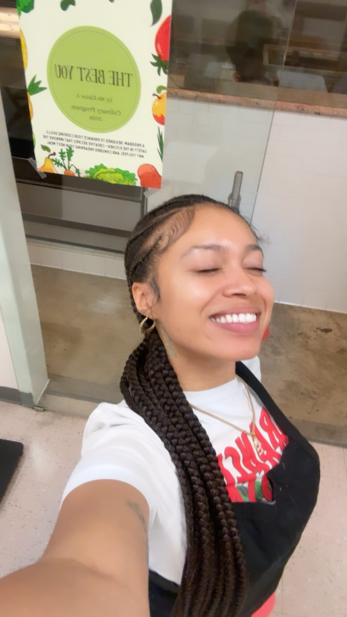 Young woman with braided hair smiling, taking a selfie indoors, with a green sign in the background.