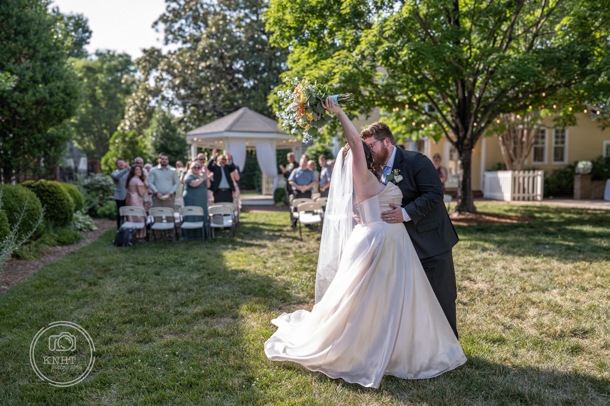 A bride and groom dance outdoors under a tree during a wedding celebration with guests in the background.