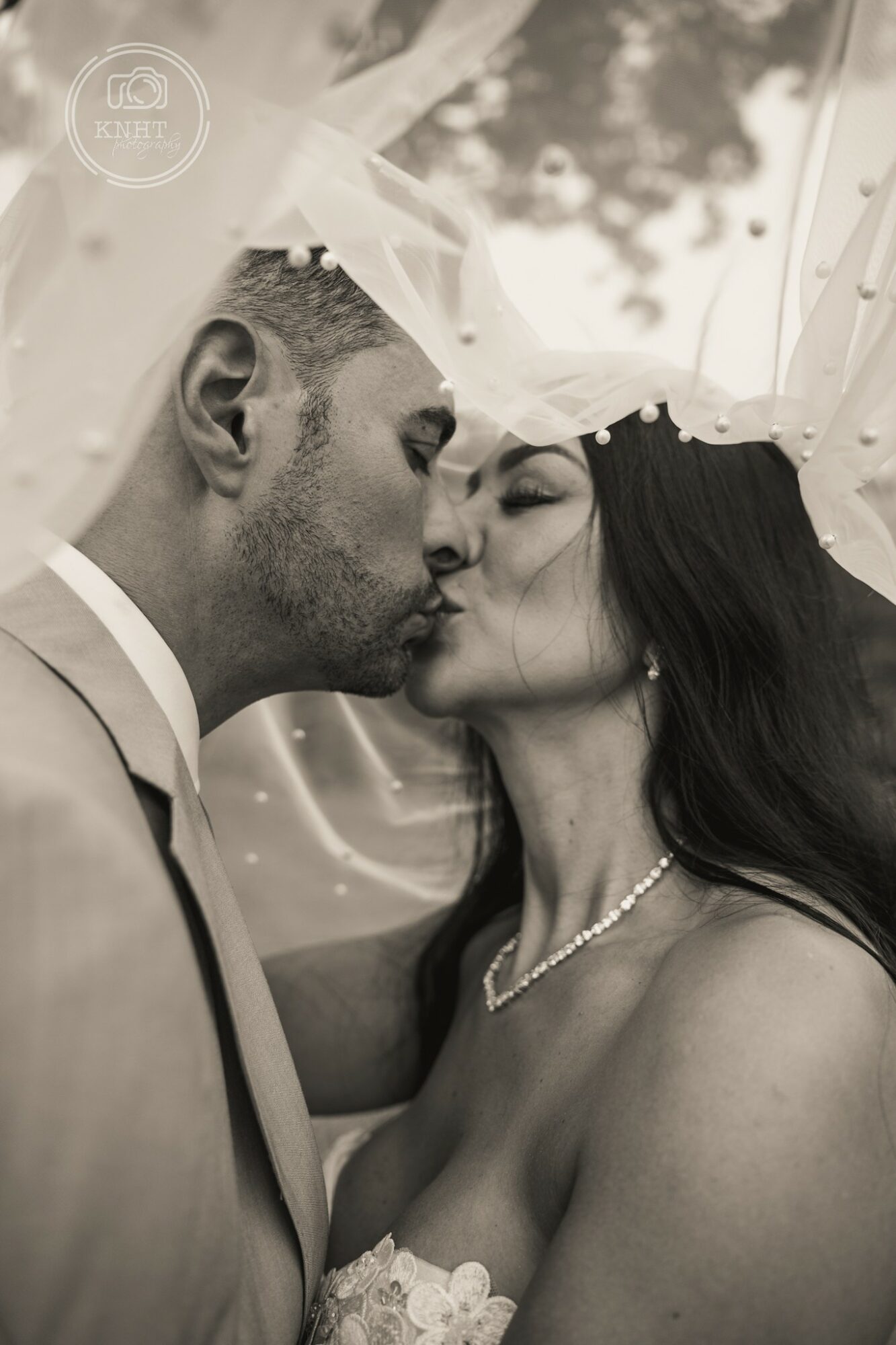 A couple kissing, dressed in wedding attire, with a veil and outdoor background, in black and white.