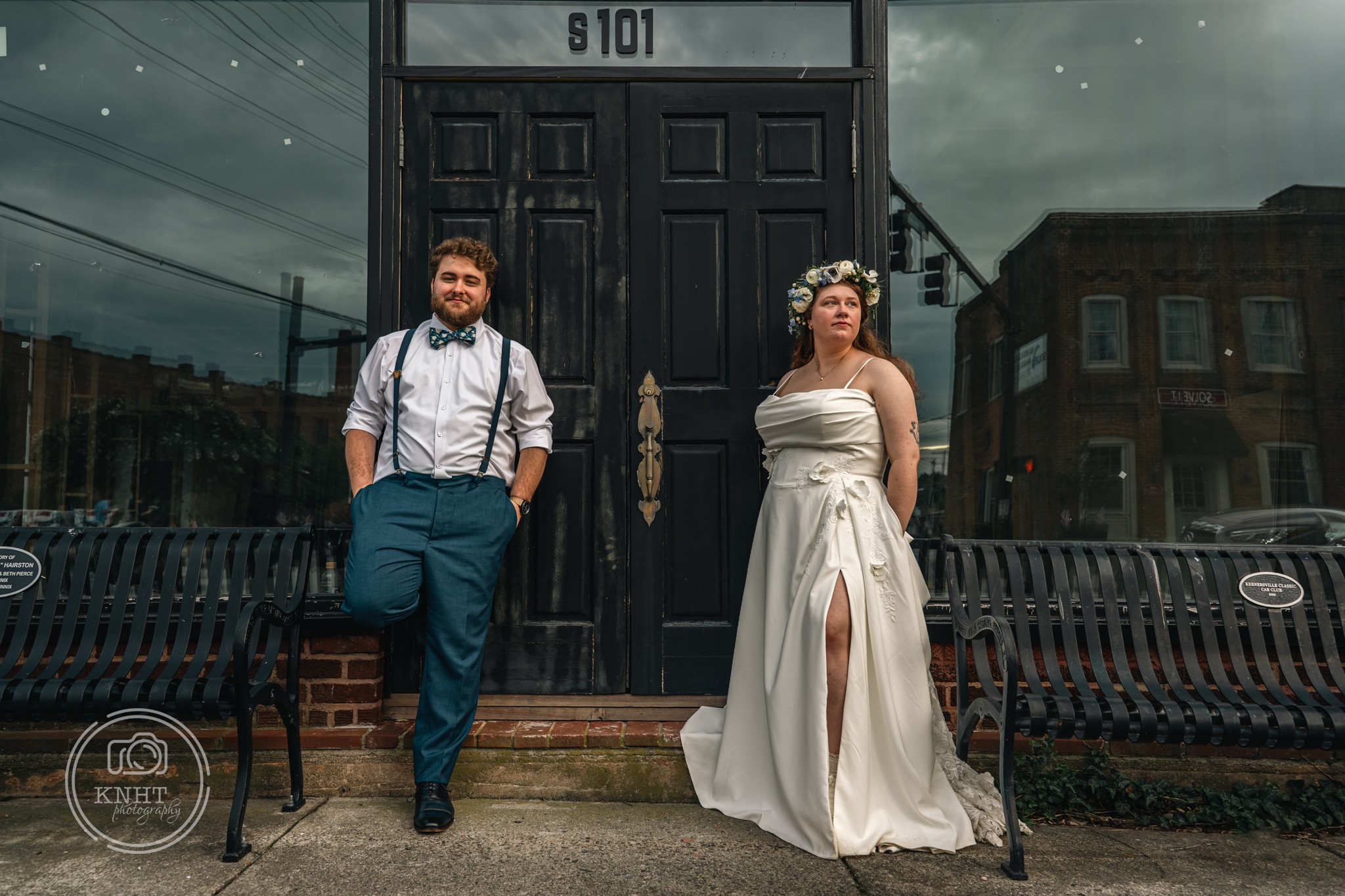 A man and woman stand outside a building with a black door, benches, and a cloudy sky in the background.