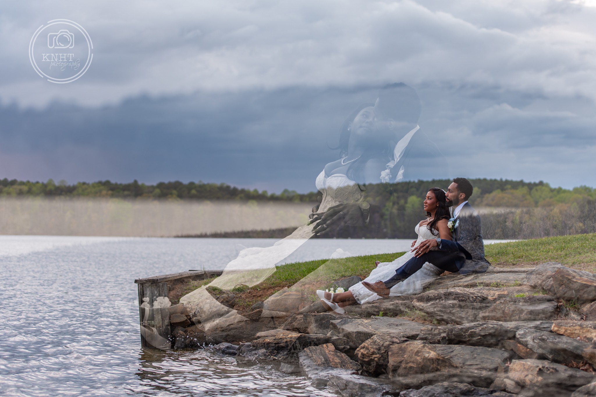 Two people sit on rocks by a lake, with a cloudy sky and distant shoreline in the background.