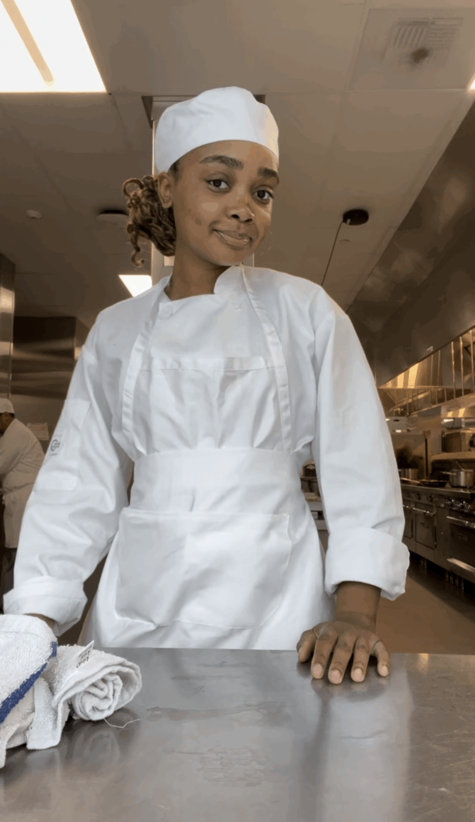 Young woman in white chef uniform and hat standing in kitchen, smiling, with hands on counter.