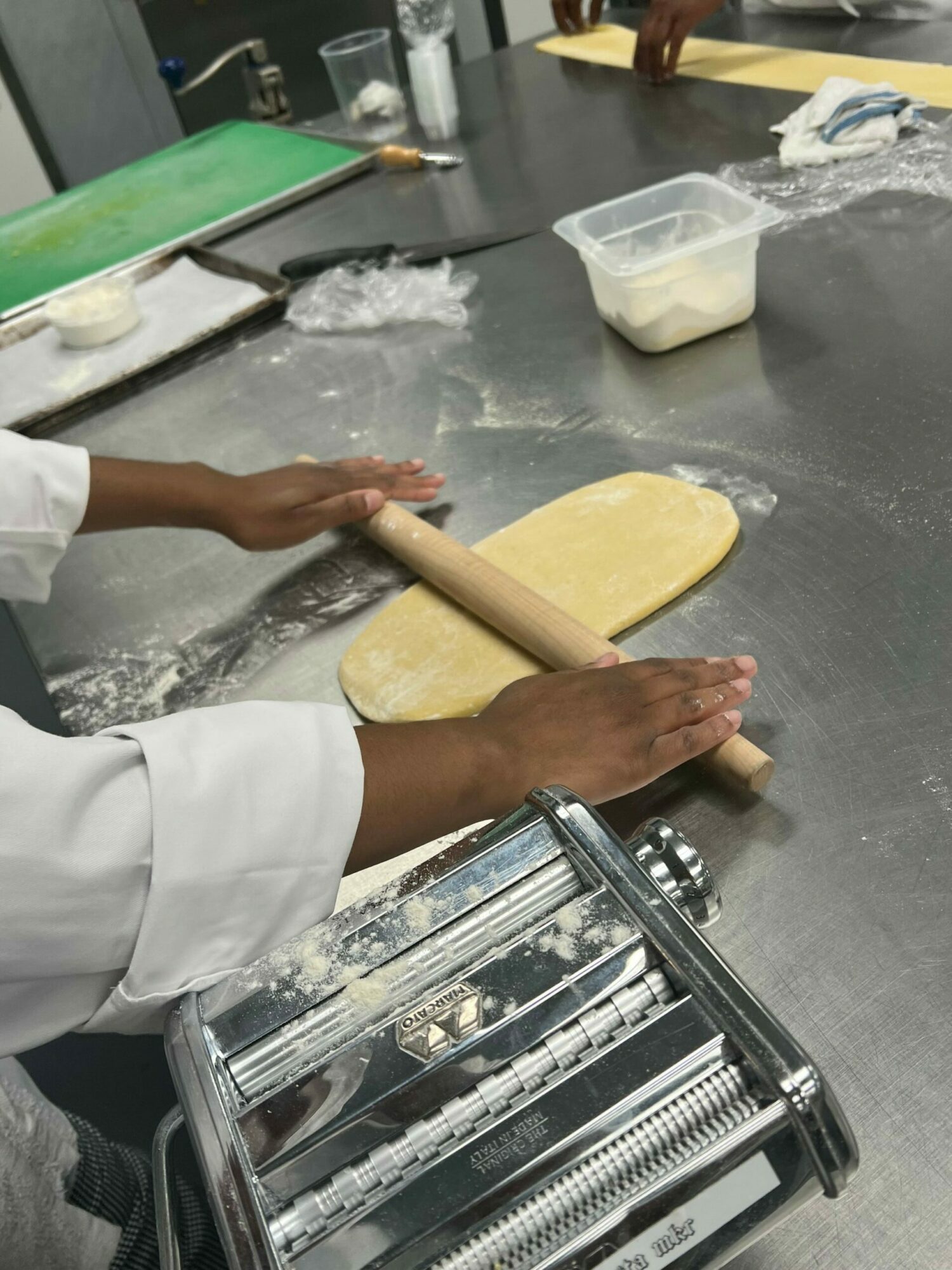 Person rolling out dough with a rolling pin on a kitchen counter, with a pasta machine nearby.