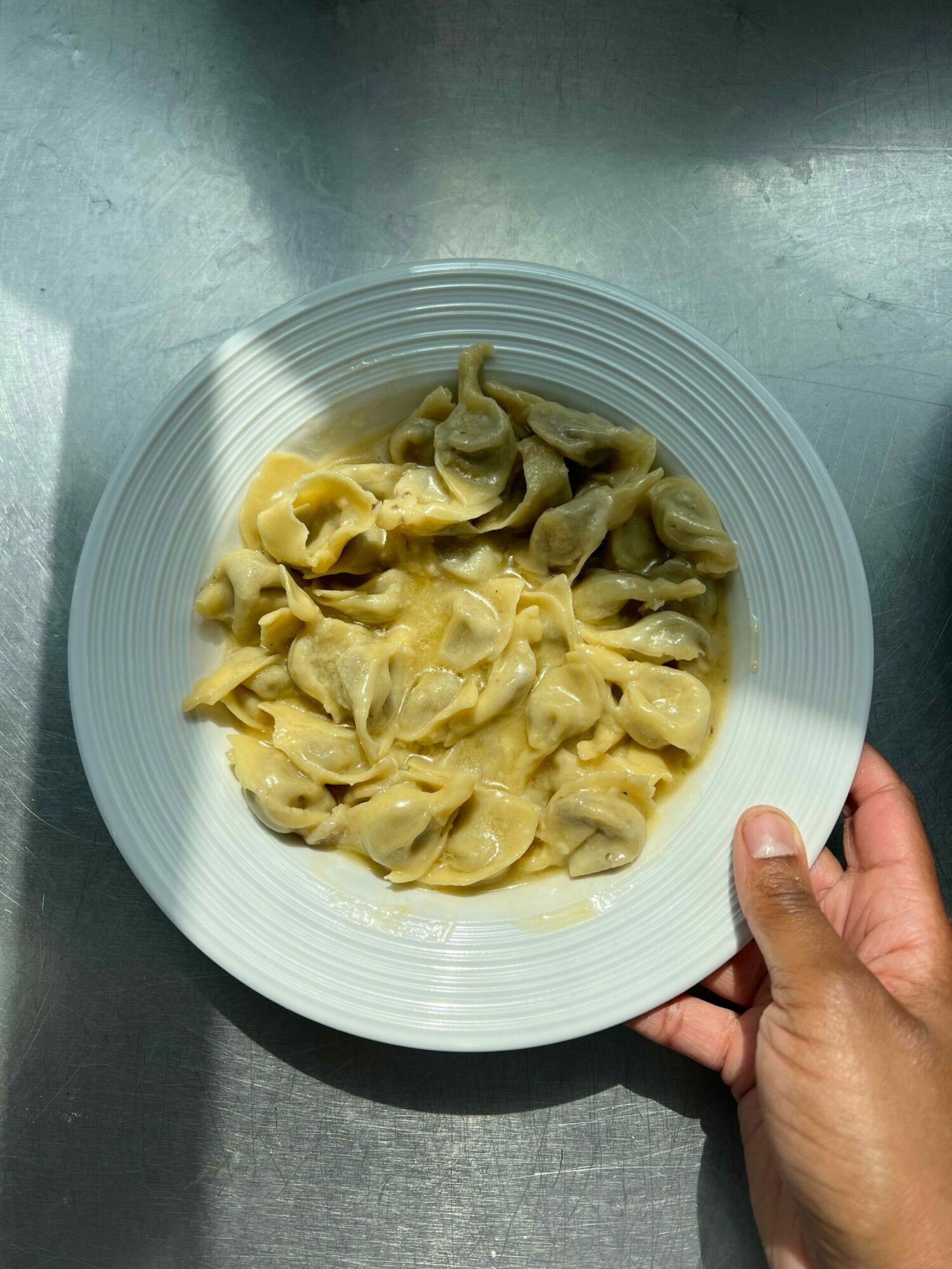 Hand holding a white bowl of cooked tortellini pasta on a gray surface.