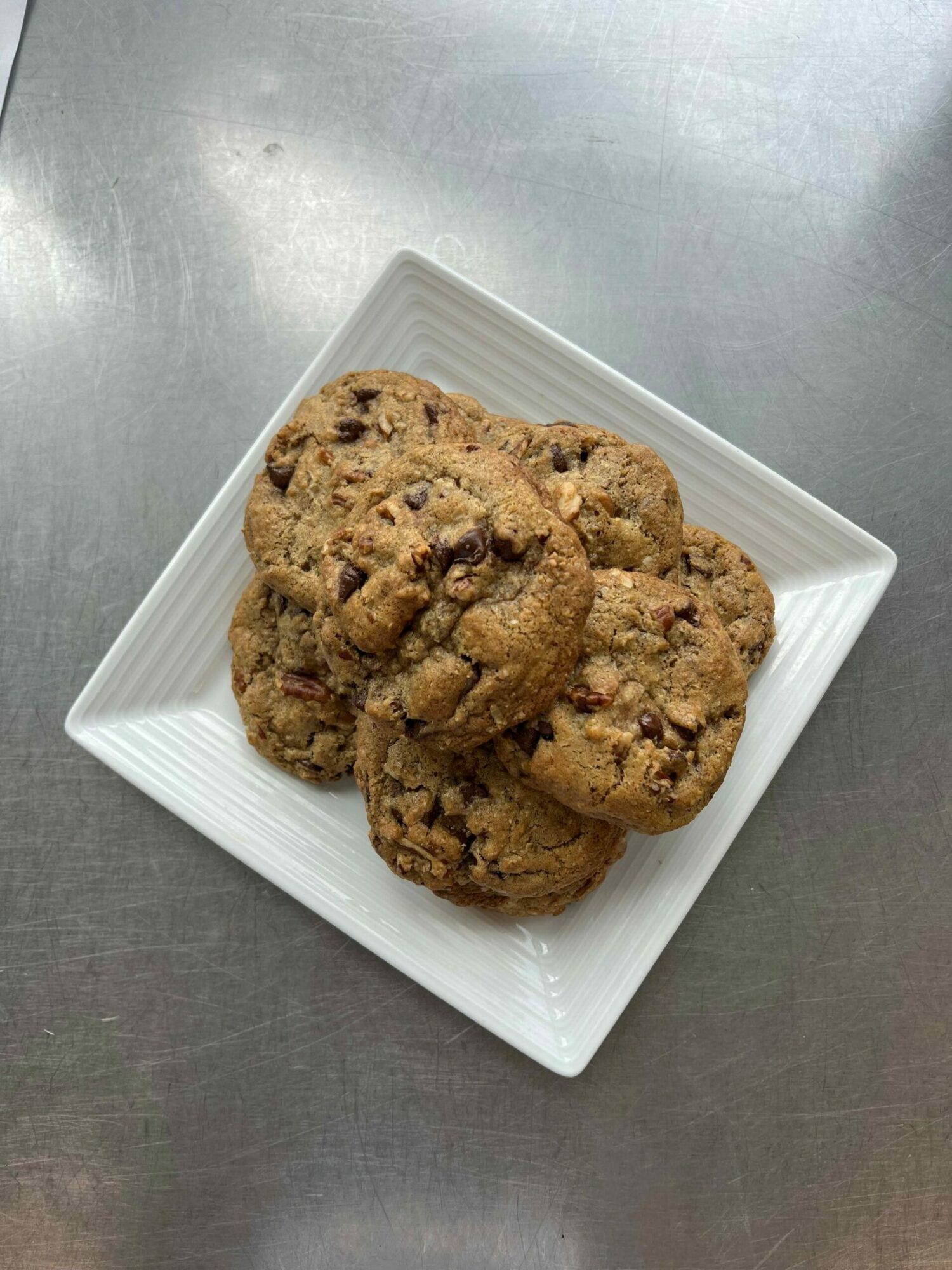 Three chocolate chip cookies on a white square plate, on a metallic surface.