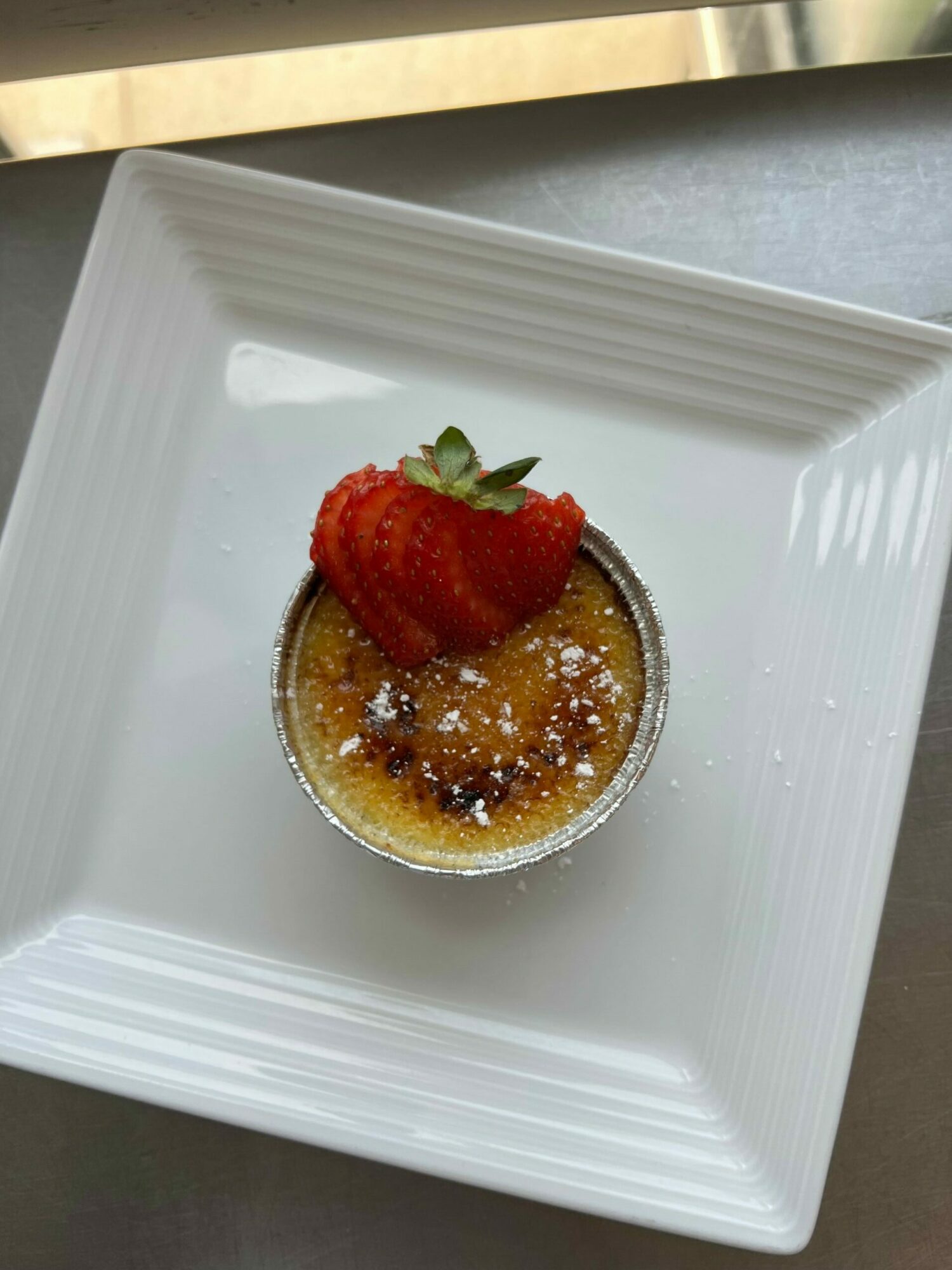 Dessert with a strawberry on top, served on a white square plate, with a caramelized sugar topping, sprinkled with powdered sugar.