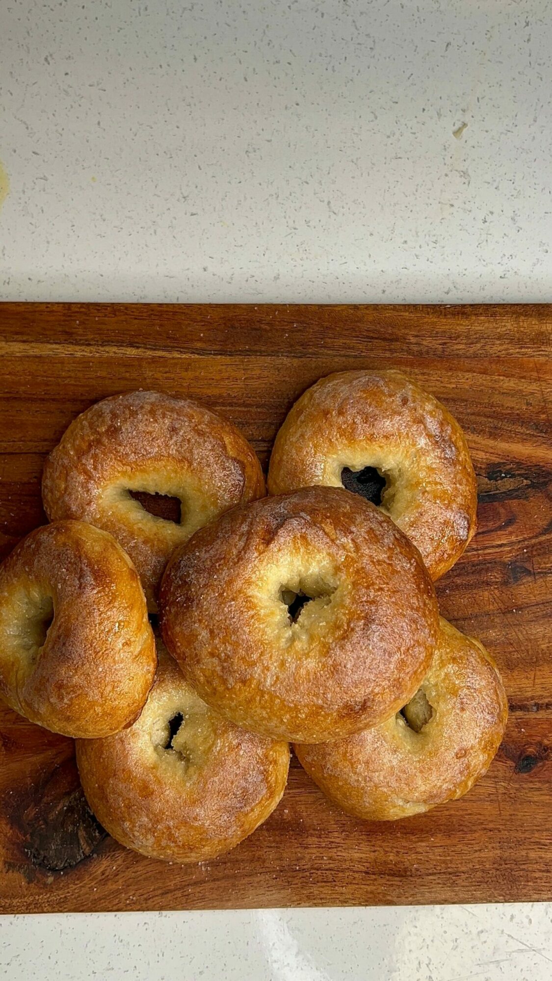 Six bagels with a hole in the center, placed on a wooden cutting board, with a light-colored surface nearby.