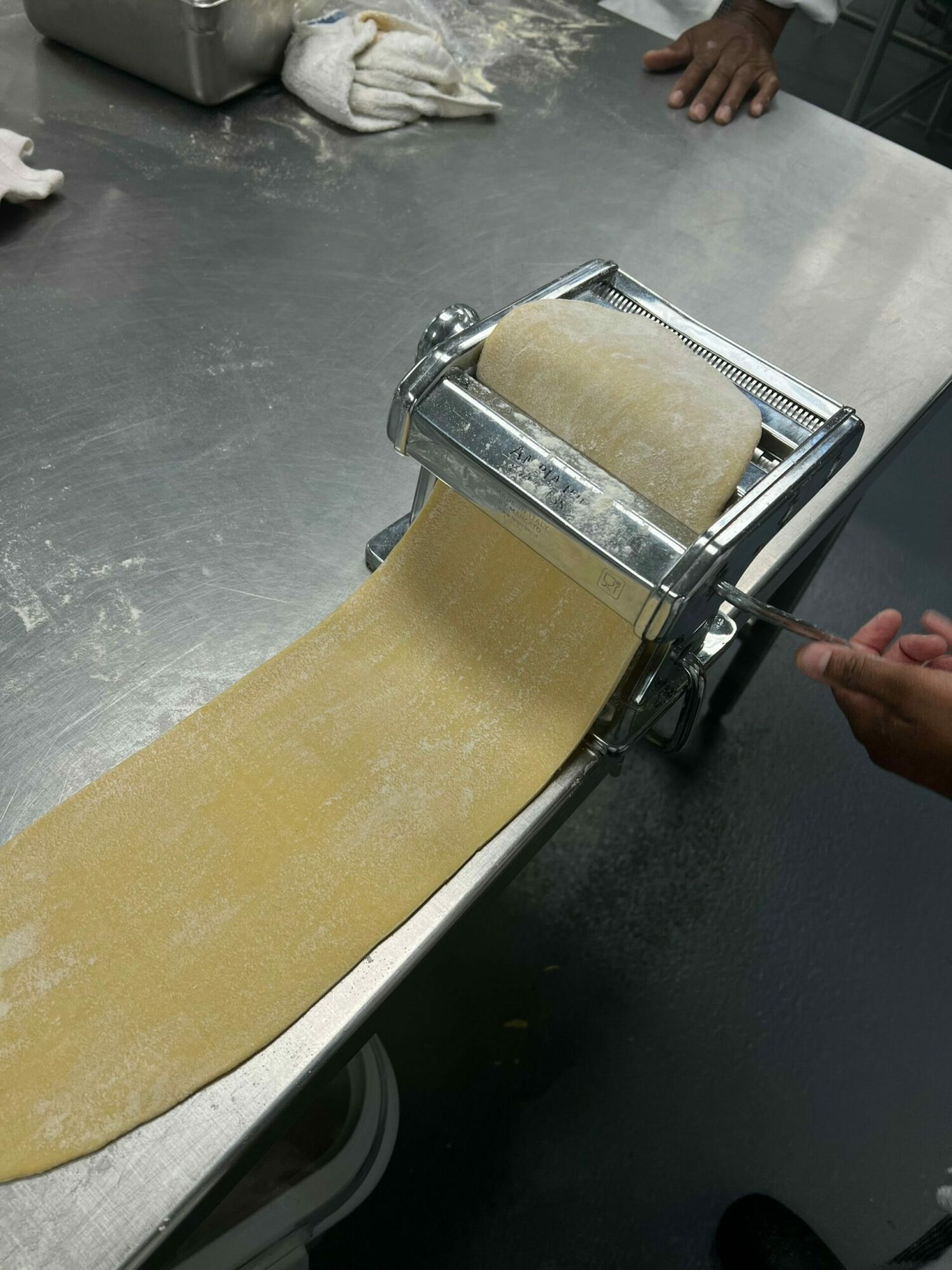 Person uses a pasta machine to roll out dough on a stainless steel surface.
