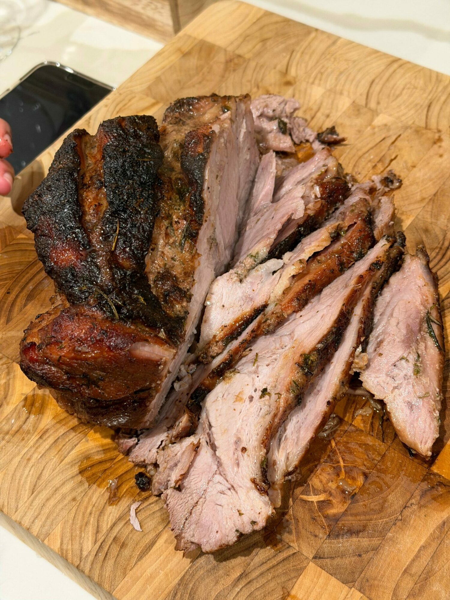 Cooked meat sliced on a wooden cutting board with a dark crust, some juices visible, and a smartphone partially visible at the bottom right.