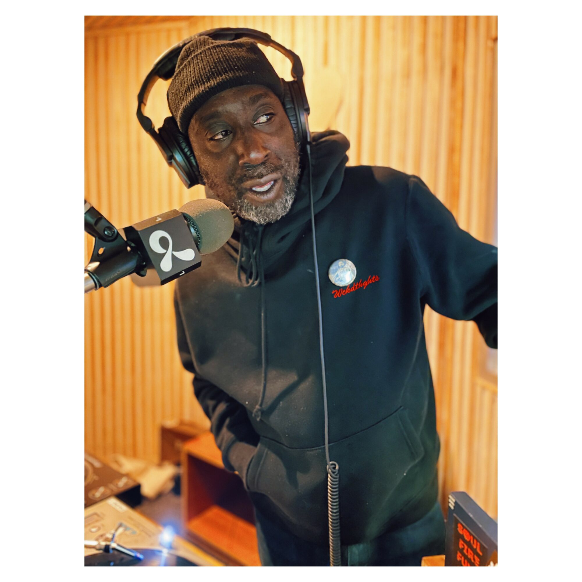 Man wearing headphones and a beanie, speaking into a microphone in a recording studio, with wooden paneling background.