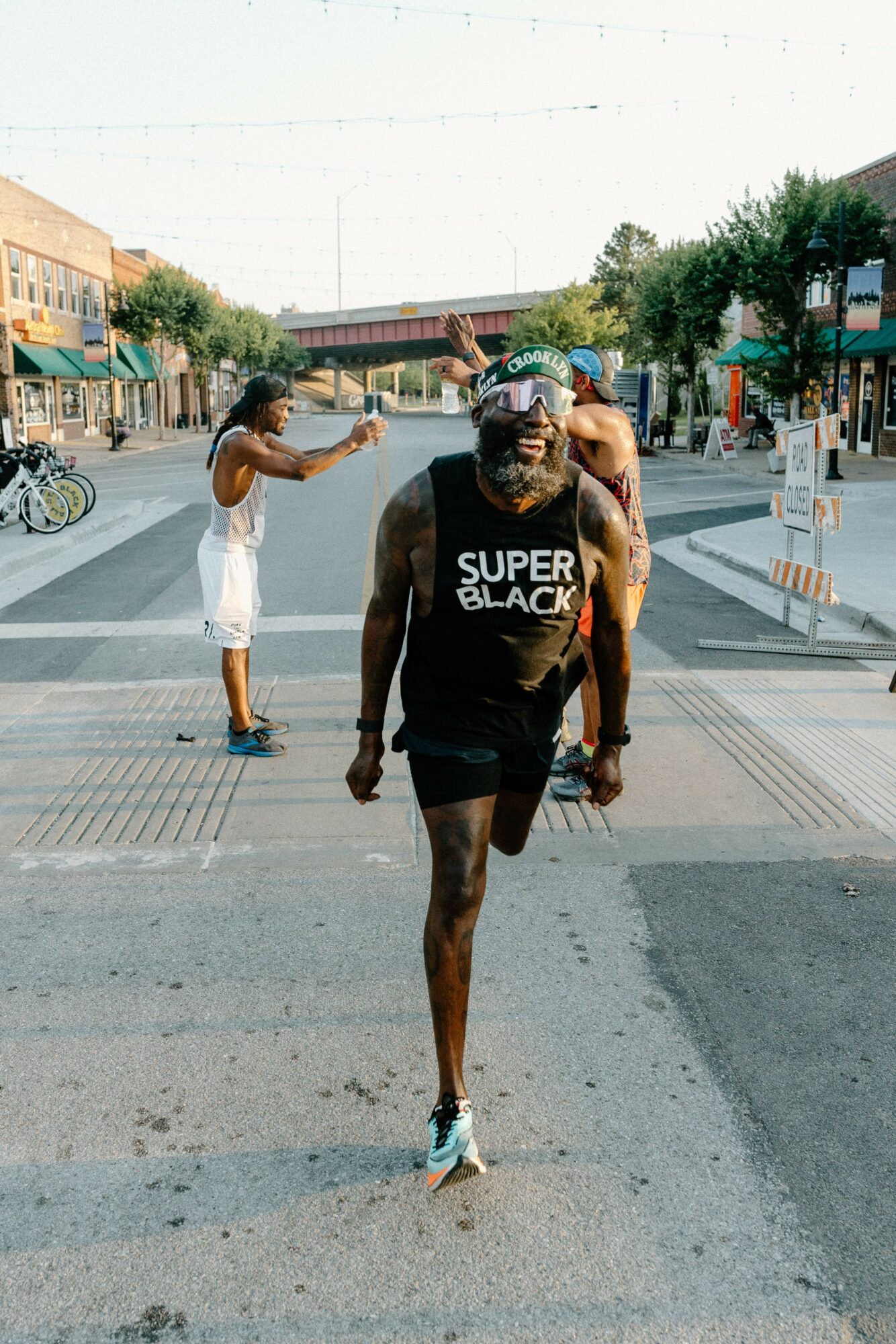 Man running on street wearing sunglasses and a tank top, with people and shops in background.