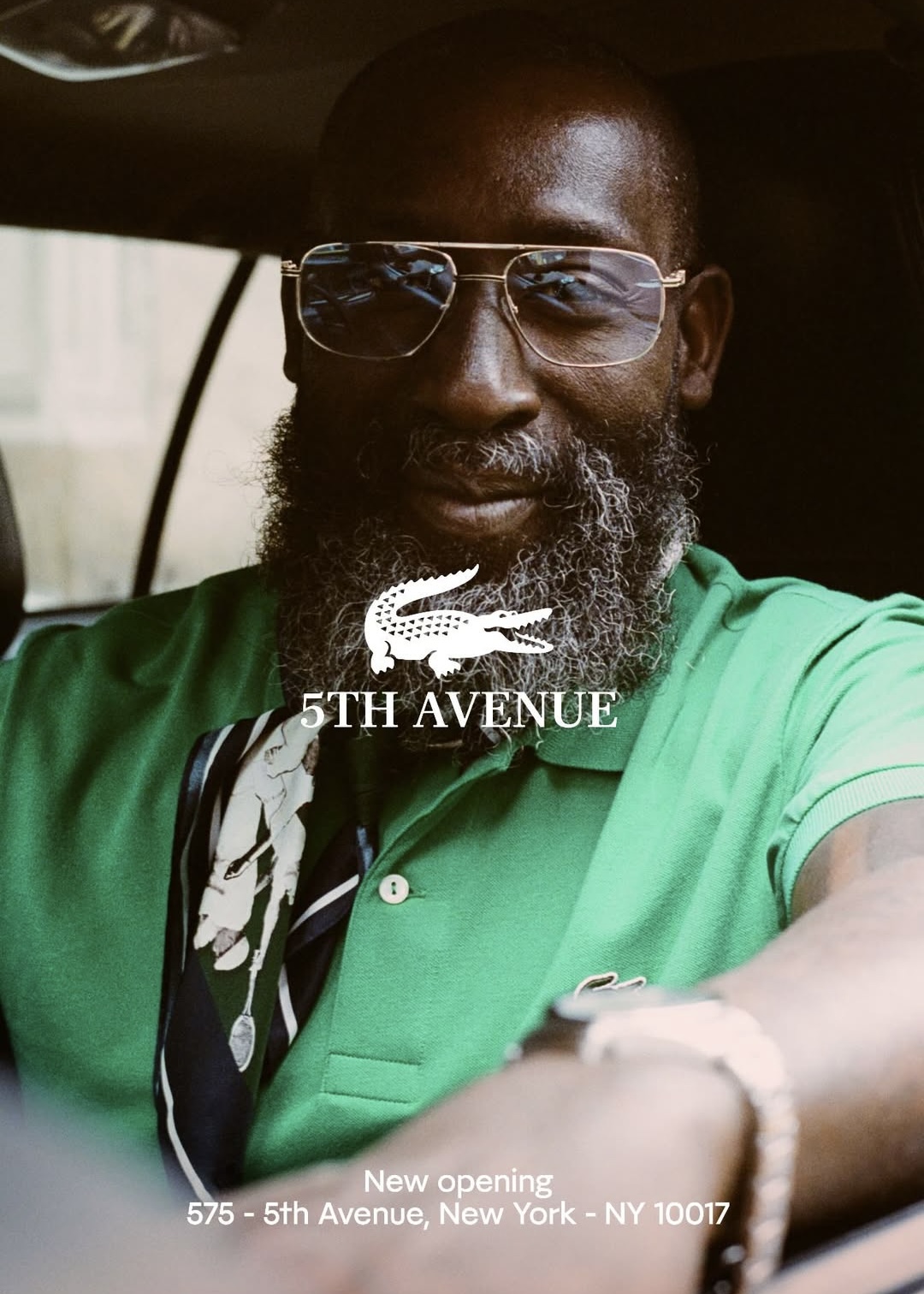 Man with glasses and beard wearing a green shirt, sitting inside a vehicle, smiling at the camera.