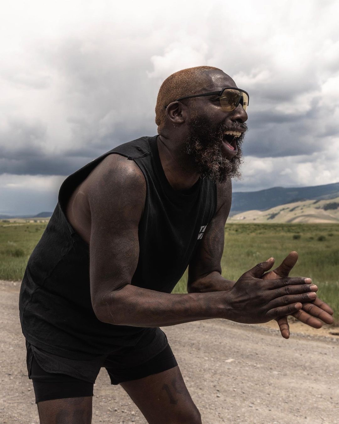 Man with glasses and beard laughing outdoors, hands clasped, cloudy sky background.