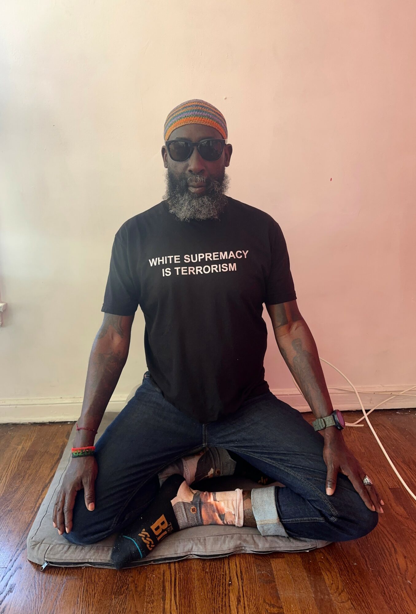 Man sitting cross-legged on a cushion, wearing sunglasses, a headband, and a black T-shirt with white text, against a plain wall.