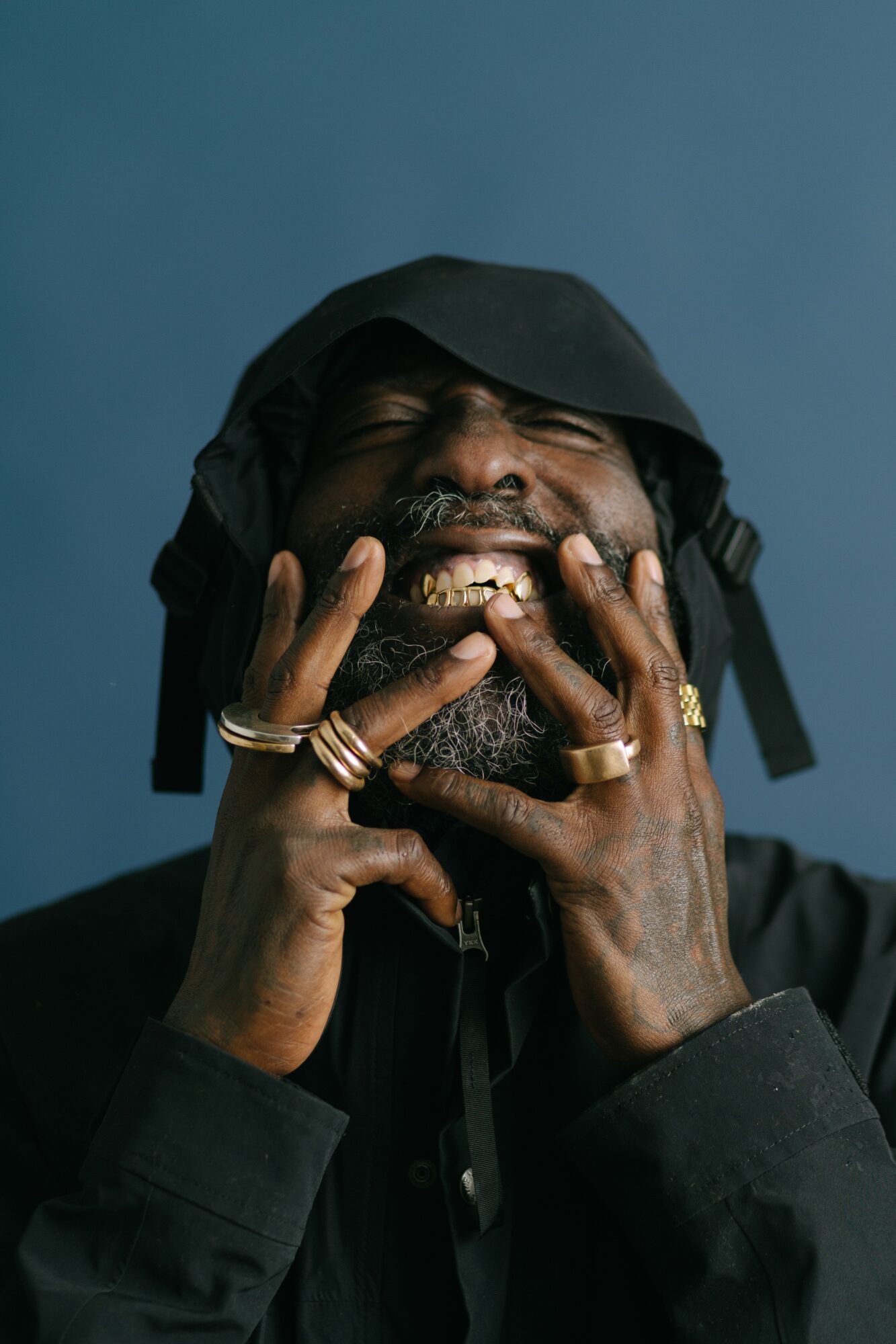 Person with a black head covering, smiling with hands near face, wearing multiple rings, against a blue background.