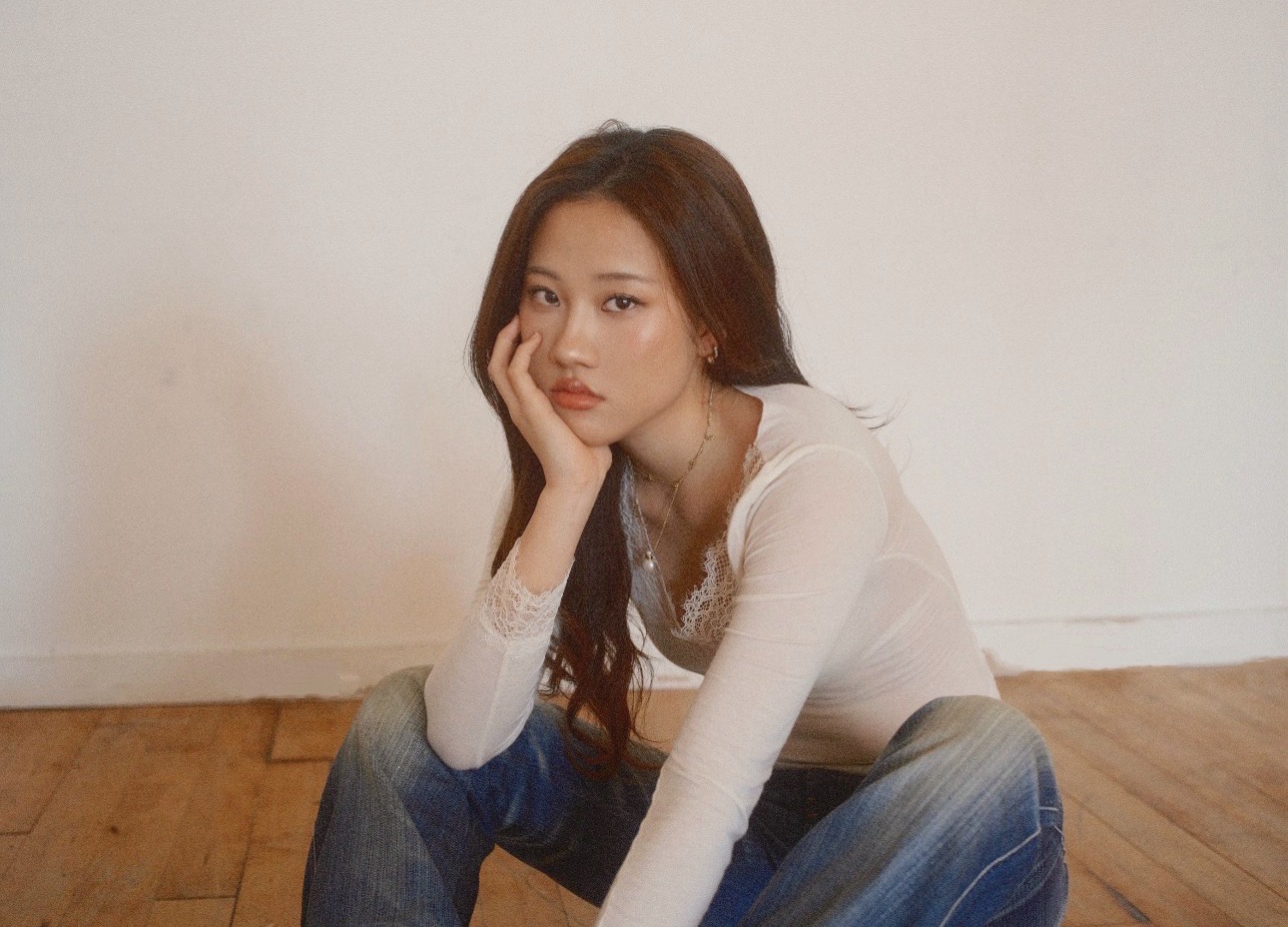Young woman with long dark hair sitting on wooden floor, resting her face on her hand, wearing a white top and jeans.