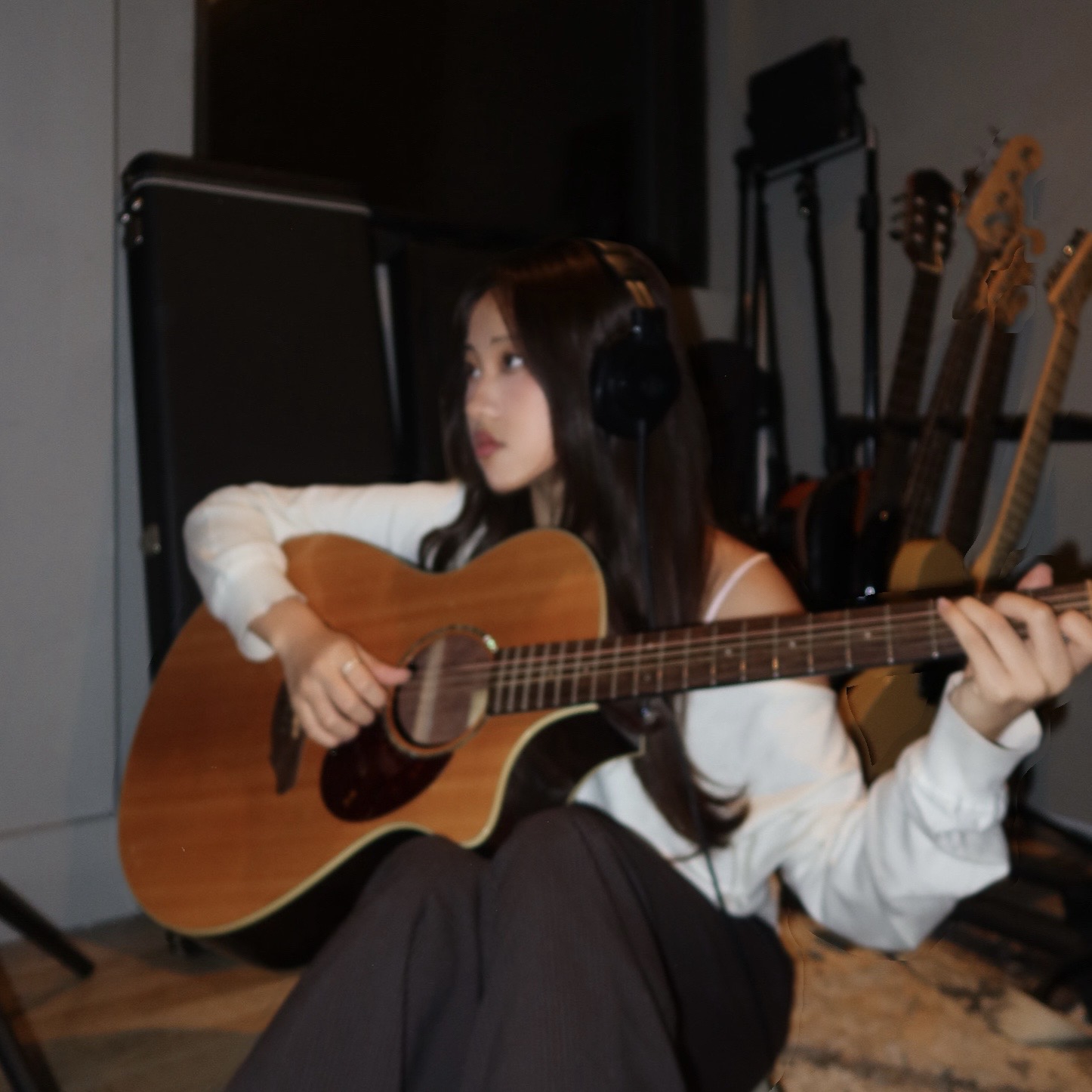 Young woman with long dark hair playing an acoustic guitar, wearing headphones, sitting indoors with guitars in the background.