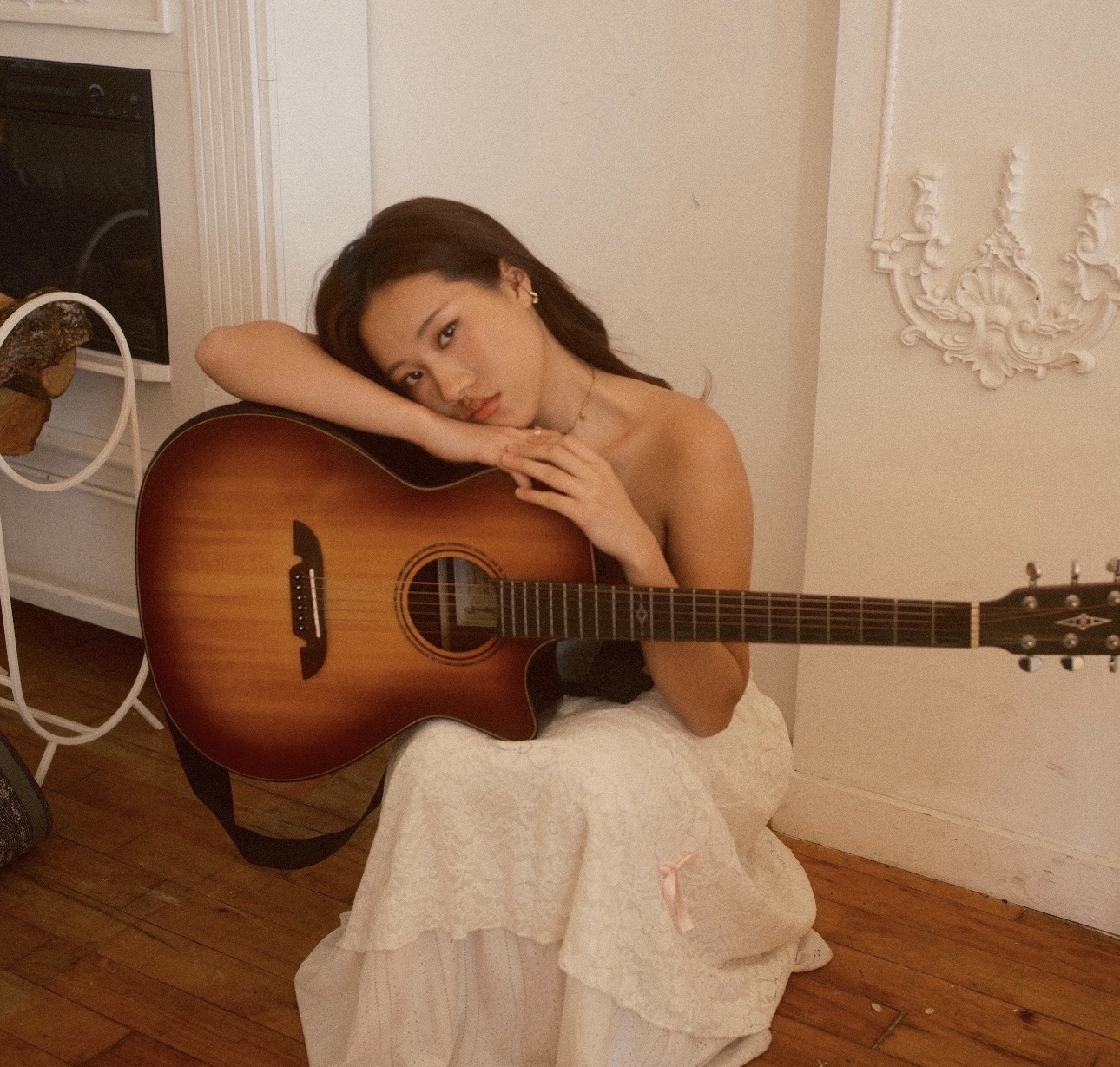 Young woman sitting on the floor with a guitar, resting her head on her arm, in a room with white walls and wooden floor.