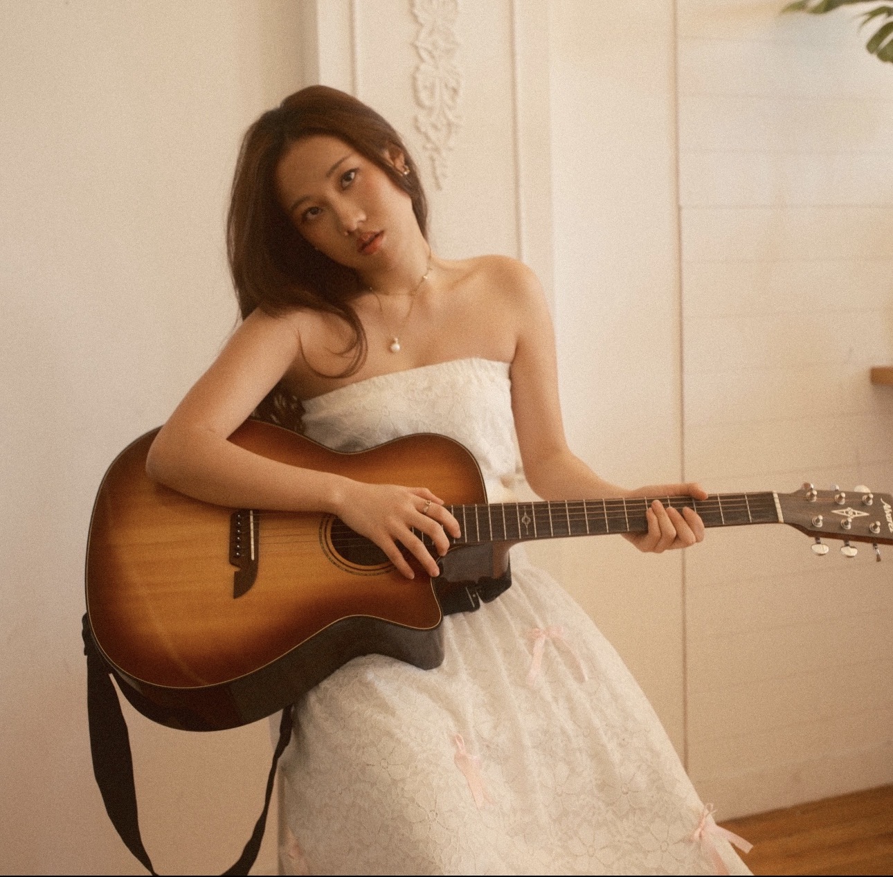 Young woman in a white dress holding an acoustic guitar indoors, looking at the camera.