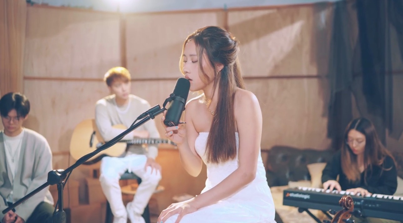 A woman singing into a microphone with three people playing instruments in the background, in a wooden room.