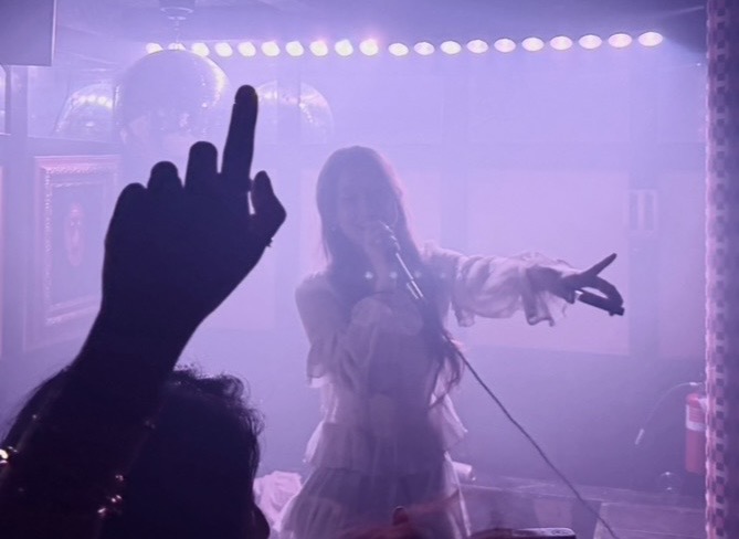 Person singing on stage with microphone, pointing, in a foggy, colorful setting, audience hand raised, dim lighting.