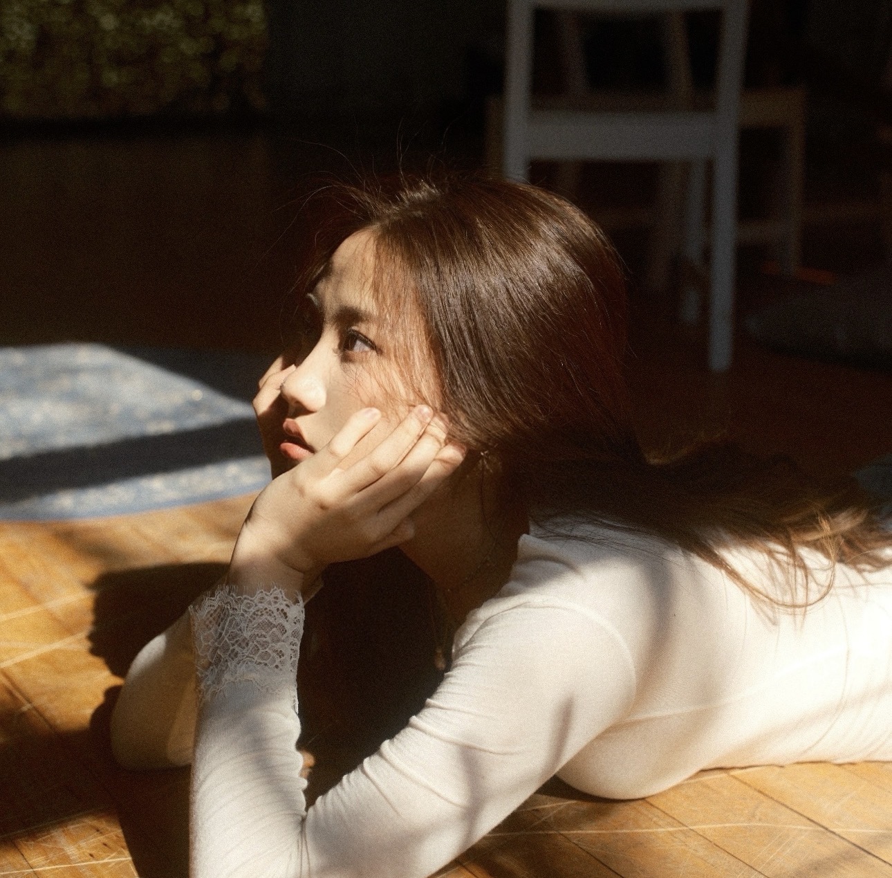 Young woman with long hair lying on a wooden surface, resting her head on her hand, looking to the side, sunlight on her face.